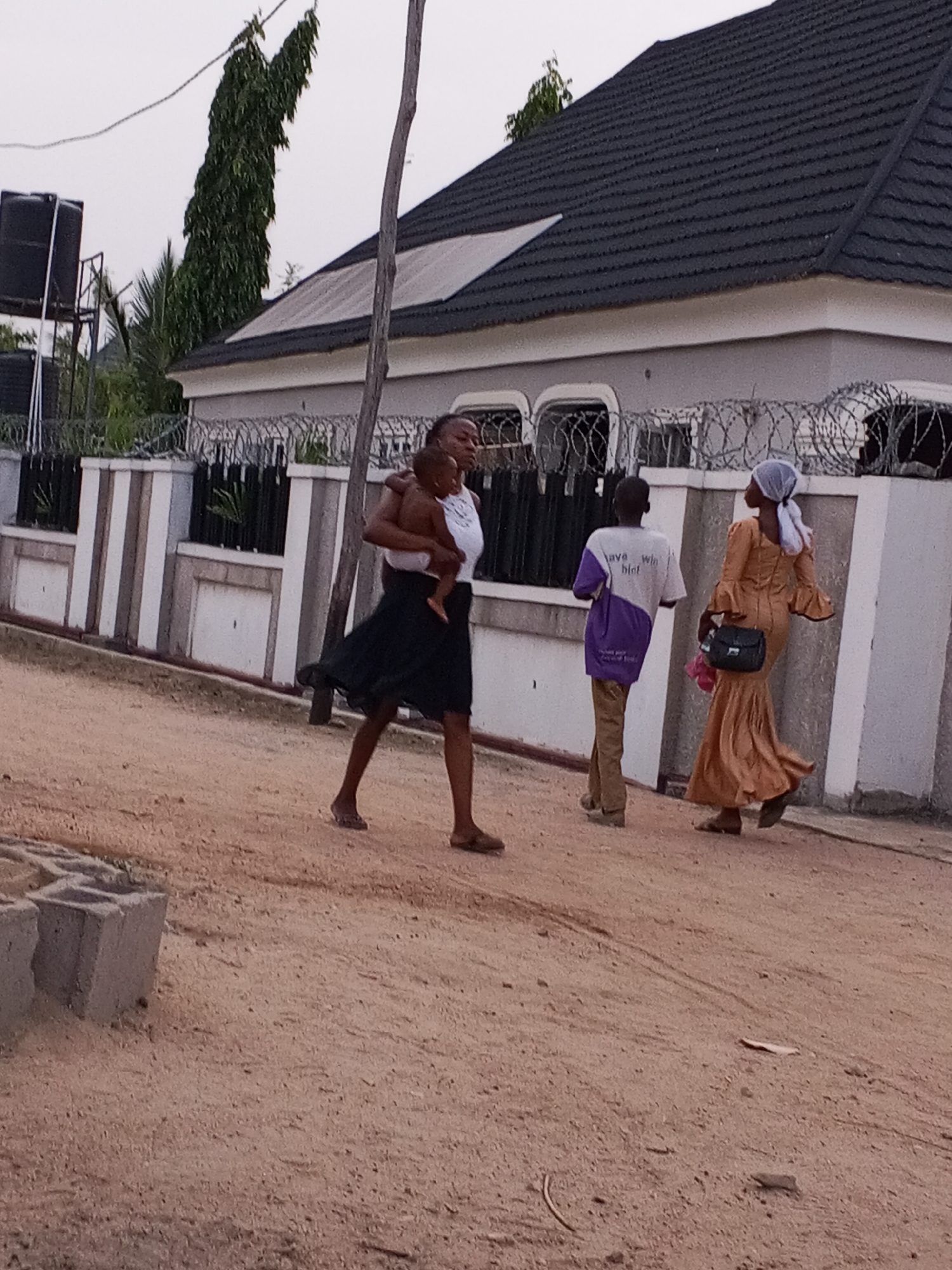The scene captures a street in Adonkolo, Nigeria. A woman carries a baby while walking on a dusty, sandy road. Behind her, two other individuals, one in a purple and white shirt and the other in a long, orange dress and a white headscarf, are also walking. The background includes a building with a dark grey roof and a fence topped with barbed wire. There are water tanks on top of the roof. A bare tree branch and a tall, thin tree are also visible near the building. In the foreground, concrete blocks are partially visible.