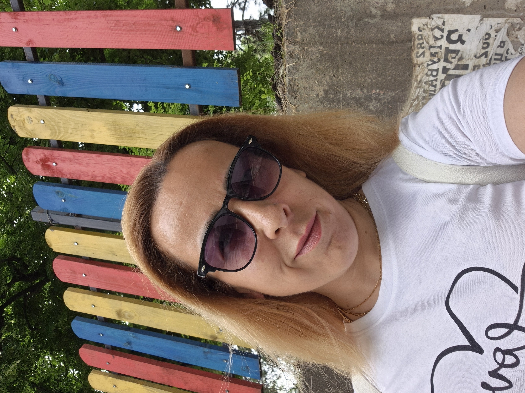 A woman is photographed sitting near a multi-colored painted bench with her face toward the camera. The bench is composed of horizontal planks painted red, yellow, and blue. The woman has light brown hair and is wearing sunglasses, a gold necklace, and a white t-shirt. There are trees visible in the background behind the bench, along with some sort of concrete and tattered paper structure to the upper right. The location is Unknown, Unknown.
