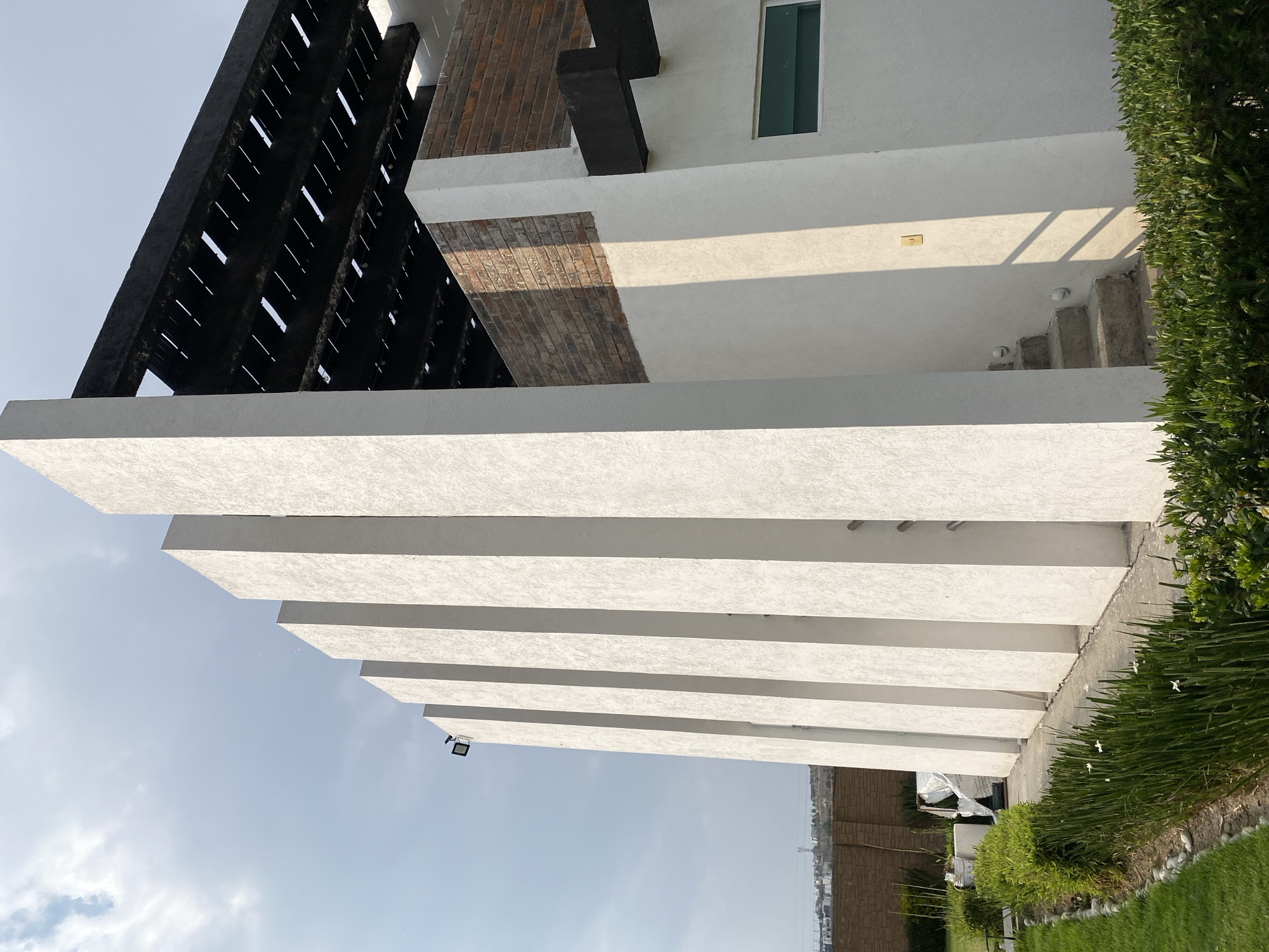 The image shows an architectural structure in Unknown, Unknown, viewed from a low angle. The structure features a series of horizontal, white concrete beams or slabs that extend outwards, casting shadows. Above these beams is a building with a black, slatted framework element and brickwork details. A single rectangular window is visible on the white portion of the building. To the right, there is greenery including a hedge or bush, and a short set of concrete steps. The sky is pale blue with some cloud cover.