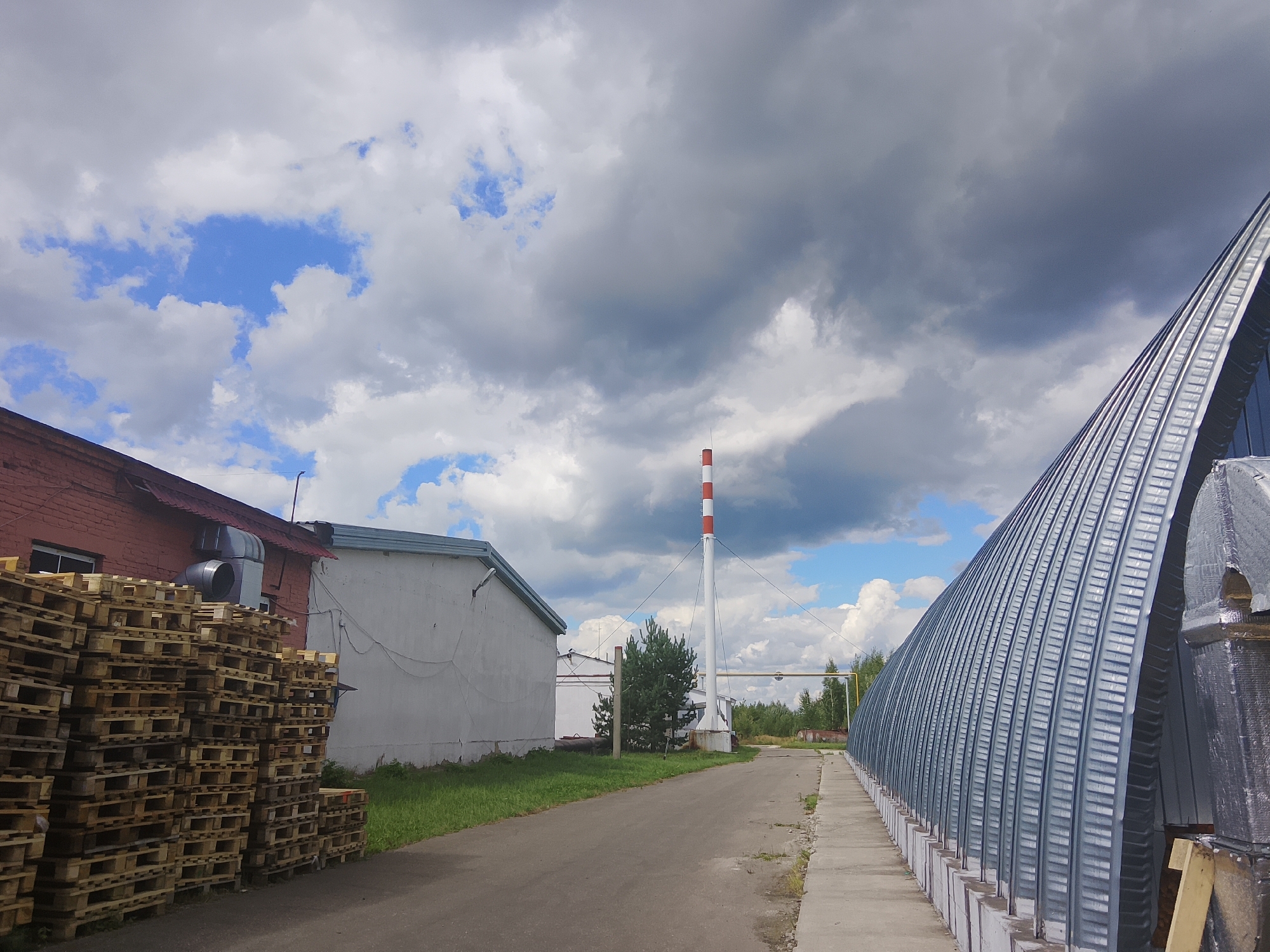 An outdoor, eye-level, wide shot captures an industrial area under a partly cloudy sky. On the left, a red brick building with a dark red roof is piled high with wooden pallets. Next to it is a white, low-slung building. In the center of the frame, a tall, slender industrial chimney with red and white stripes rises against the sky. A paved road runs through the scene, bordered by grass and trees in the distance. On the right, a large, arched metal structure with a ribbed silver exterior dominates the foreground. The overall impression is one of an active, albeit somewhat sparse, industrial facility.