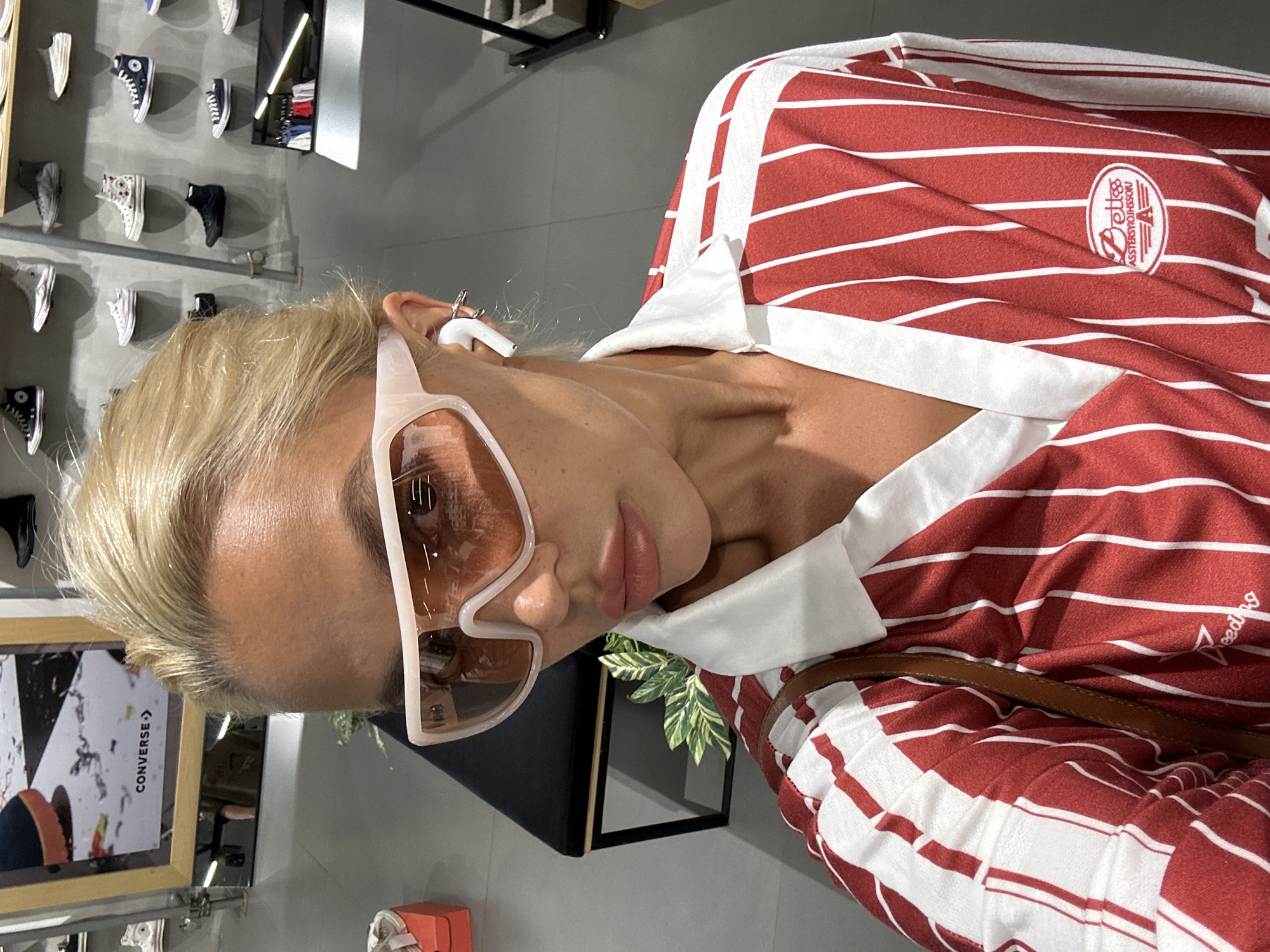 The image shows a woman with blonde hair and sunglasses, wearing a red and white striped shirt, indoors, in a store. In the background, there's a display of Converse shoes and a framed poster, suggesting the location might be a Converse store. The shirt has a logo on the upper right side. It is not possible to determine if the location is Kuta, Indonesia from the image alone.