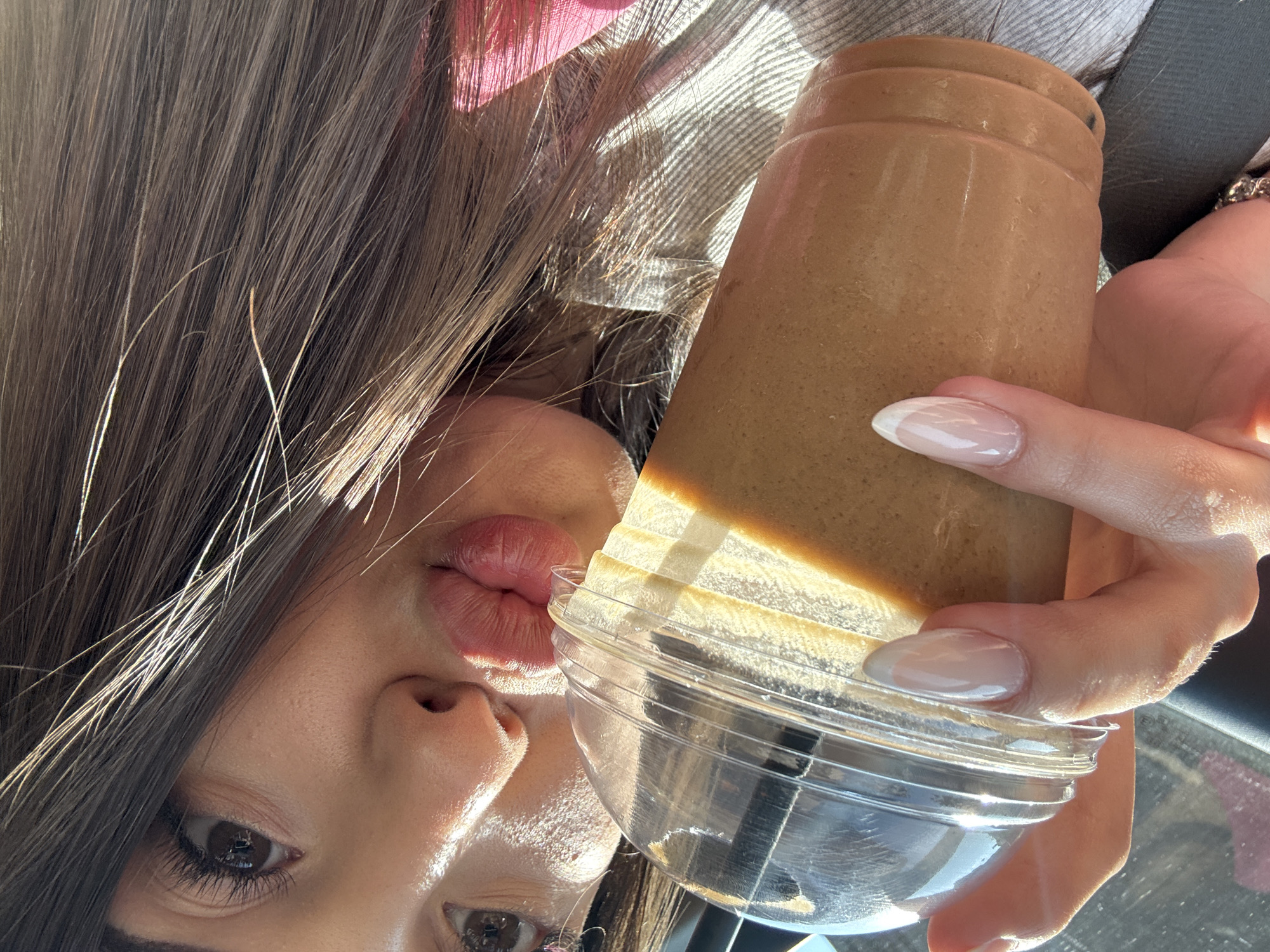 The image depicts a female individual situated inside a vehicle in the city of Rakovski, Bulgaria. She is holding a clear plastic cup containing a light brown beverage, which is approximately two-thirds full. The cup is fitted with a transparent domed lid and contains a black straw. Her left hand, adorned with a silver-toned ring on the ring finger, grasps the cup. Her nails are manicured in an almond shape with a classic French tip design. The female's face is partially visible, oriented towards the viewer. Her lips are puckered in a "kiss" expression, and her right eye is open, with extended eyelashes. Dark brown hair, illuminated by strong sunlight causing bright highlights, flows across her shoulder and partially obscures the right side of her face. Her skin exhibits visible texture under the direct sunlight. A dark grey seatbelt is present across her chest, indicating a car interior. She appears to be wearing a light-colored, textured garment, with a glimpse of pink material visible on her right side.