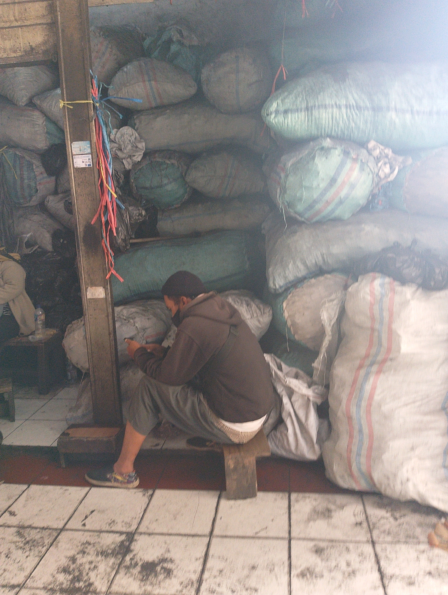 In Lembang, Indonesia, a man wearing a dark beanie and a brown hooded sweatshirt sits on a low wooden stool in front of a large stack of burlap sacks filled with what appears to be produce or other goods. He is holding a smartphone in his hands and looking down at it. The sacks are of various sizes and colors, with some featuring horizontal stripes. The scene takes place in what appears to be a market or storage area, with a tiled floor and a wooden support beam visible to the left of the man. A few colorful plastic ribbons hang from the beam. The lighting is somewhat hazy, possibly due to dust or smoke in the air.