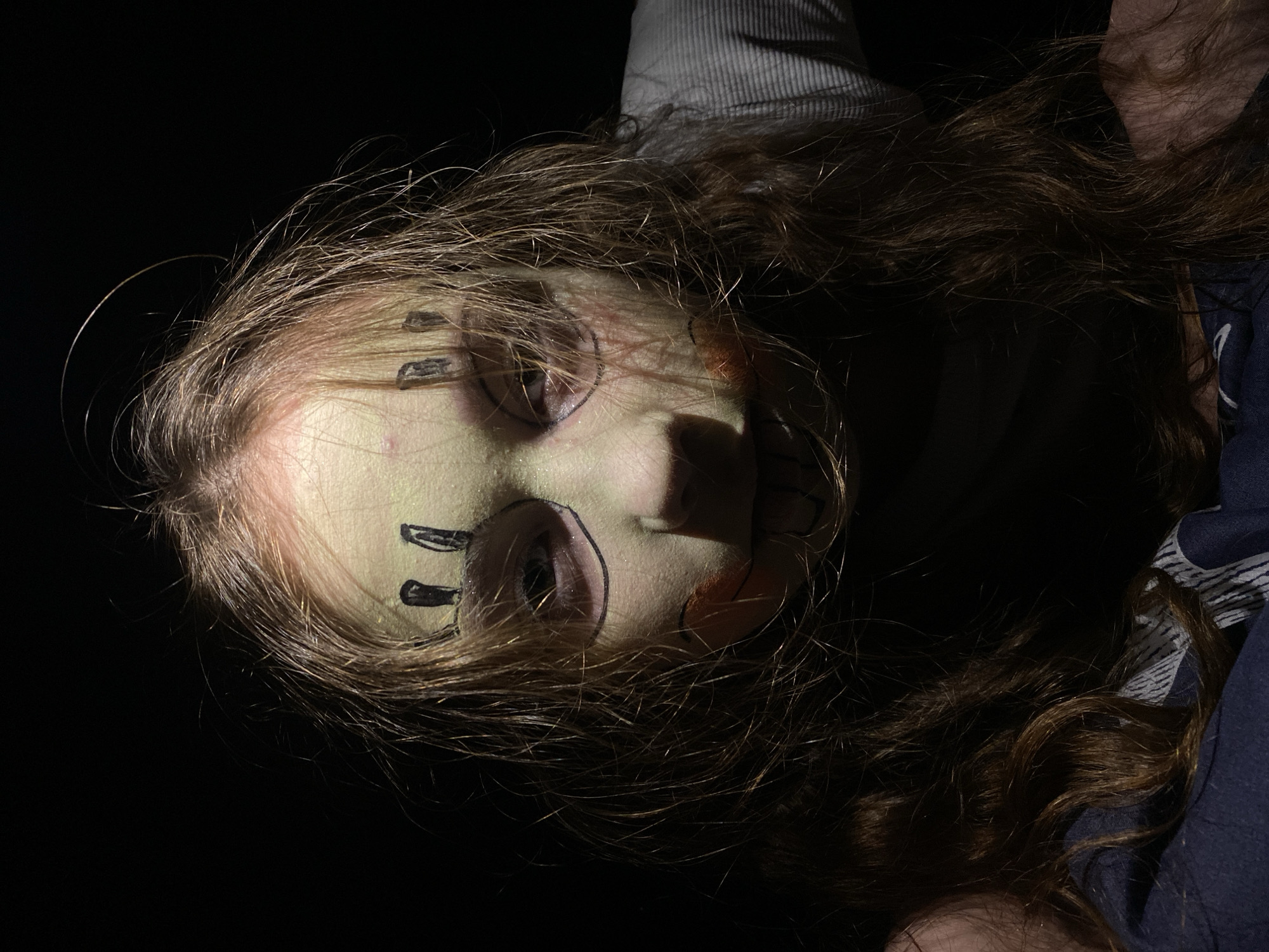 A low-angle, poorly lit portrait depicts a person with long, light brown hair and pale green skin, possibly due to makeup or lighting. The person's face is adorned with black markings around the eyes and on the forehead, resembling tribal or theatrical makeup. The background is dark and indistinct, with a hint of clothing visible on the periphery, including a light-colored striped garment. The location is Unknown, Unknown.