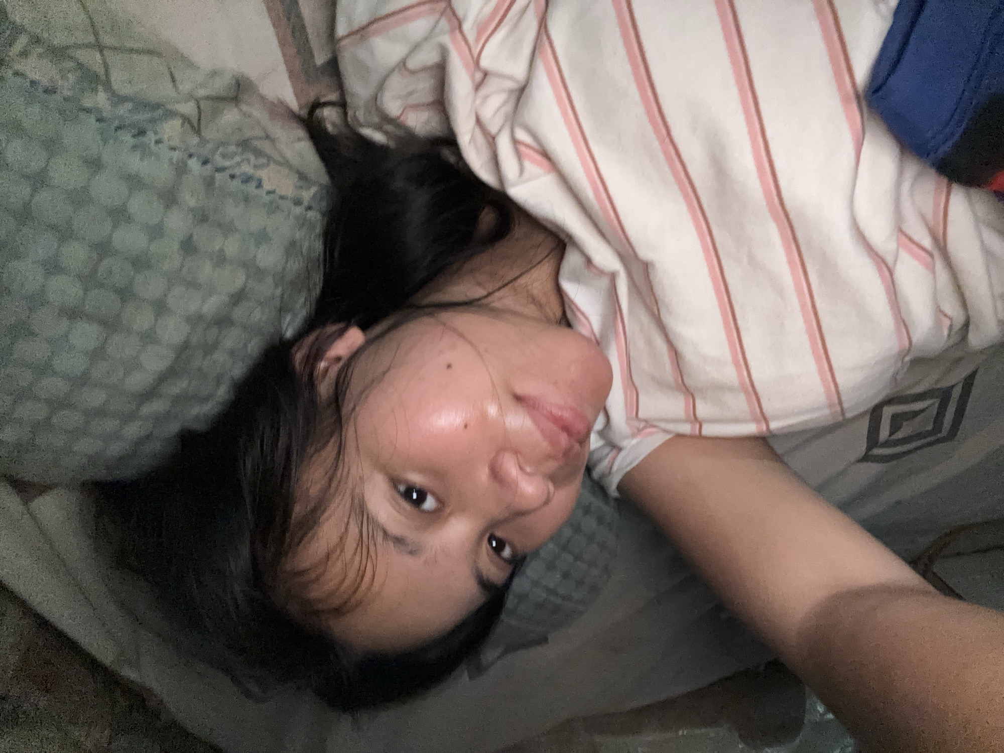 A woman lies in bed, propped up slightly on her left arm and pillow. Her eyes are open, and she is looking at the camera. She is wearing a white and pink striped shirt. The room appears to be dimly lit, with shadows cast on the wall behind her. The scene is consistent with a private bedroom, likely in a residential setting within East Jakarta, Indonesia.