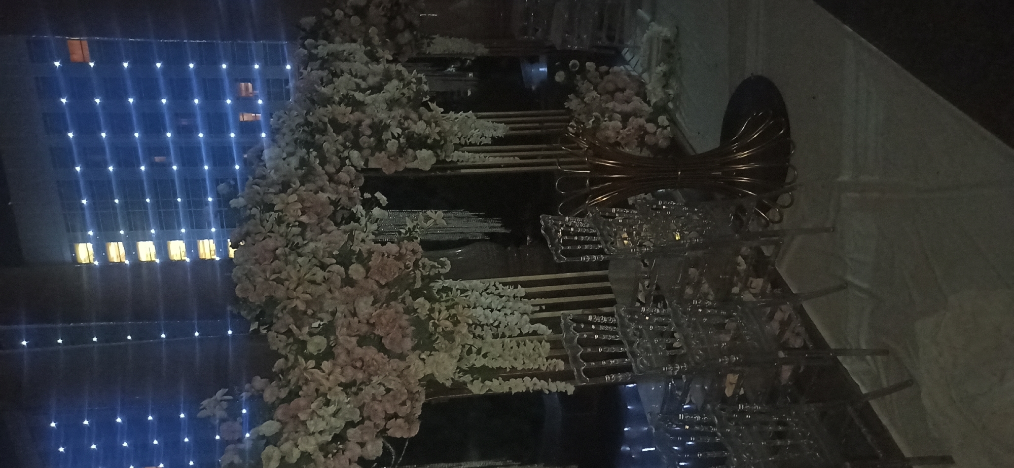The image depicts an outdoor event setup at night in Dushanbe, Tajikistan. A floral archway, adorned with light pink and white flowers and hanging white floral chains, forms the focal point. Several clear acrylic chairs are arranged in front of the archway, with some appearing to be set at tables, although the tables themselves are mostly obscured. A decorative metallic centerpiece, resembling a large, open vase with curved metallic elements, stands to the right of the chairs. In the background, a tall building is visible, illuminated by rows of white lights that create a grid pattern on its facade. The overall scene suggests a celebratory or event atmosphere, likely a wedding or formal gathering.