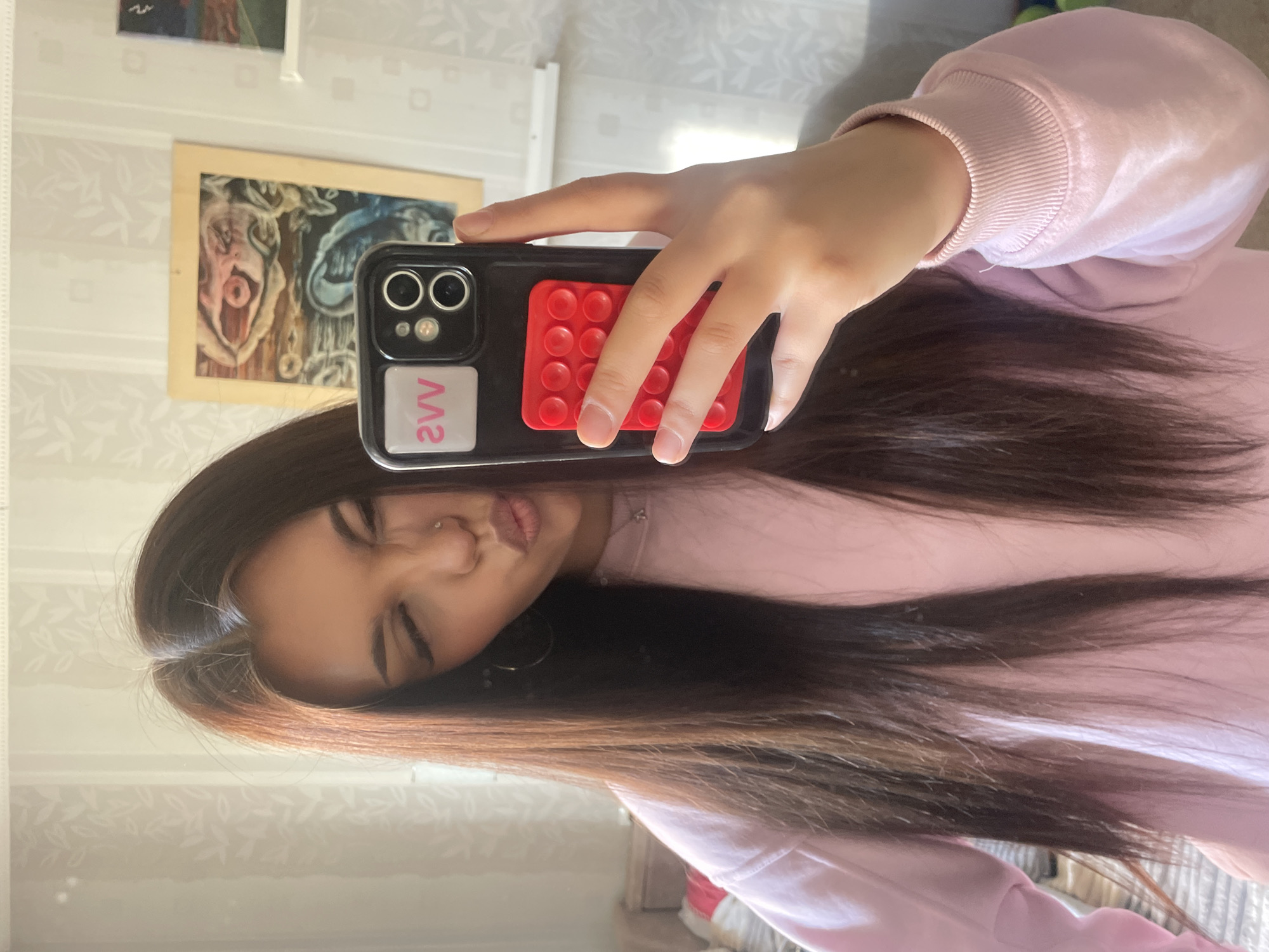 A selfie photograph depicts a young woman with long, dark brown hair and light brown highlights, wearing a light pink hooded sweatshirt. Her face is partially obscured by a black smartphone in a black case with a red, textured rectangle and a white square with "WV" printed in pink. She is making a duck-face expression. The background is a wall with light, patterned wallpaper, and two framed pictures are partially visible. The location appears to be an interior space, possibly a bedroom or apartment, in an unknown city.