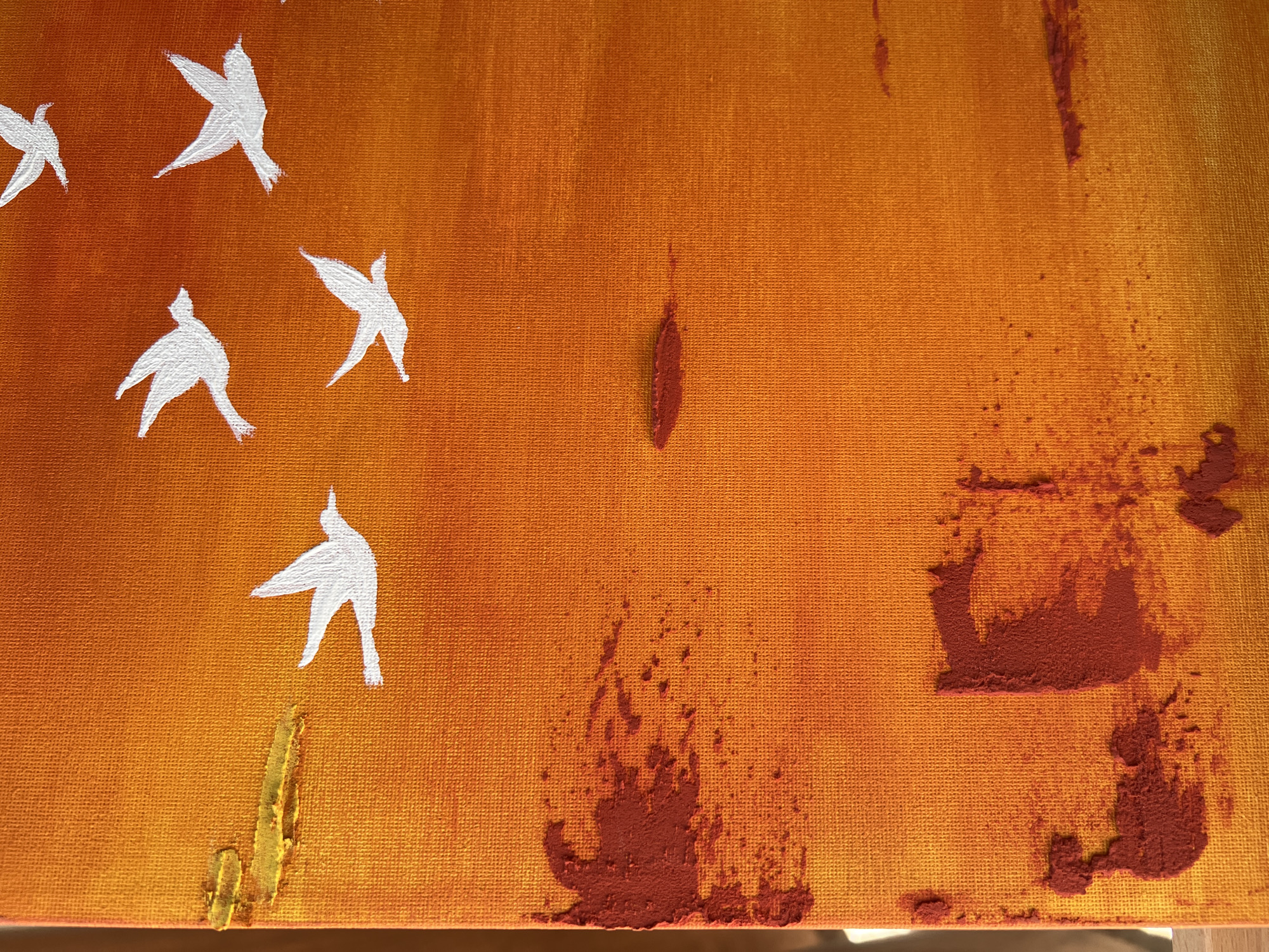 The image displays a close-up of an abstract painting with a textured canvas. The background is a gradient of warm colors, transitioning from a bright orange at the top to a deeper, more reddish-orange towards the bottom. Scattered across the canvas are stylized white silhouettes of birds in flight. Some of these birds are more distinct than others, with visible wing shapes and body outlines. In the lower left quadrant of the painting, there are patches of dark red pigment, applied in a textured, almost clumpy manner, creating a contrast with the smoother background and the white birds. There are also smaller red splatters and streaks in this area. To the left of the red patches, a few horizontal strokes of bright yellow paint are visible, appearing as textured daubs. Further to the right, some horizontal streaks of red pigment are present, appearing more subdued. The overall impression is one of a dynamic, abstract landscape, possibly representing a sunset or a flock of birds against a vibrant sky. The image does not depict any identifiable landmarks or features related to the city of Namangan City, Uzbekistan.