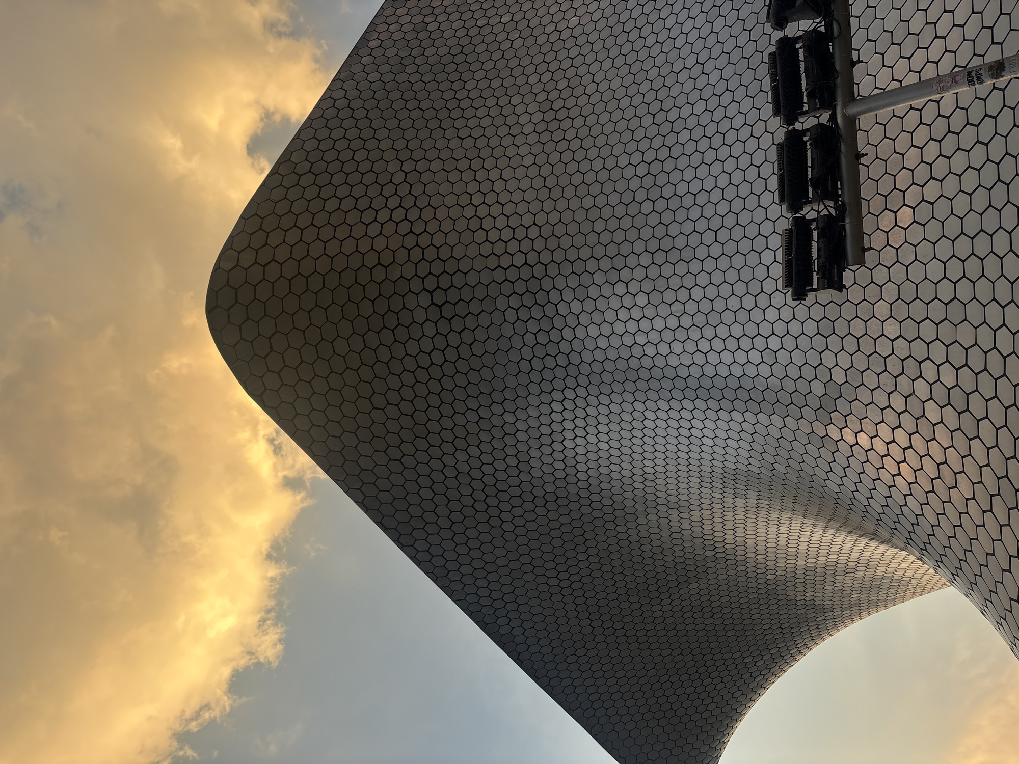 This is a low-angle shot of the Soumaya Museum in Mexico City, Mexico. The museum's exterior is covered in hexagonal aluminum tiles. The sky is visible on the left side of the frame, with golden clouds. On the right side of the frame, there is a metal pole with lights attached to it. The architecture style and design are modern.