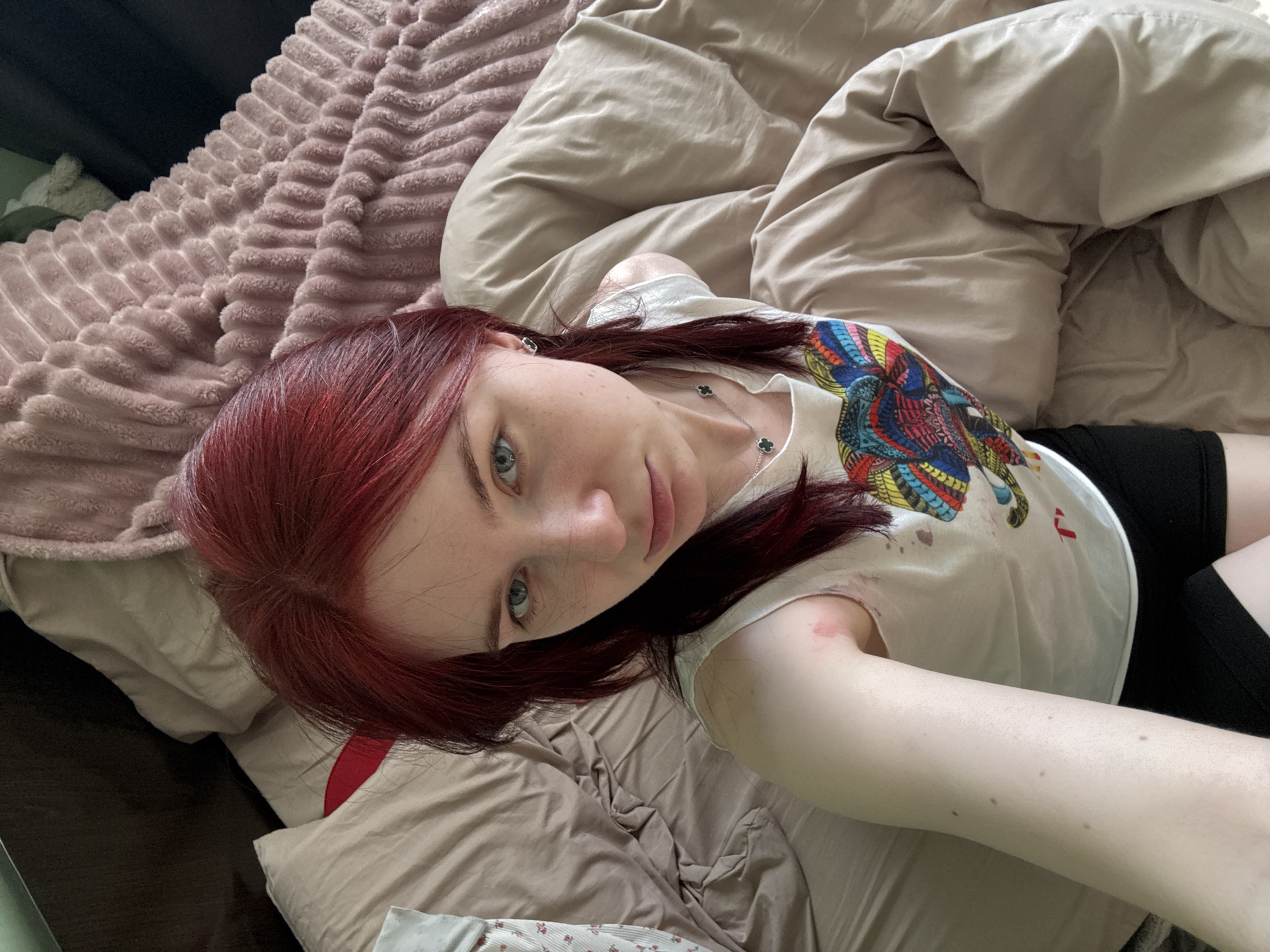 A woman with red hair is lying on a bed, looking up at the camera. She is wearing a white t-shirt with a colorful graphic on it, black shorts, and a silver necklace. Her left arm is resting on the bed. The bed has beige and pink blankets. The background is out of focus.