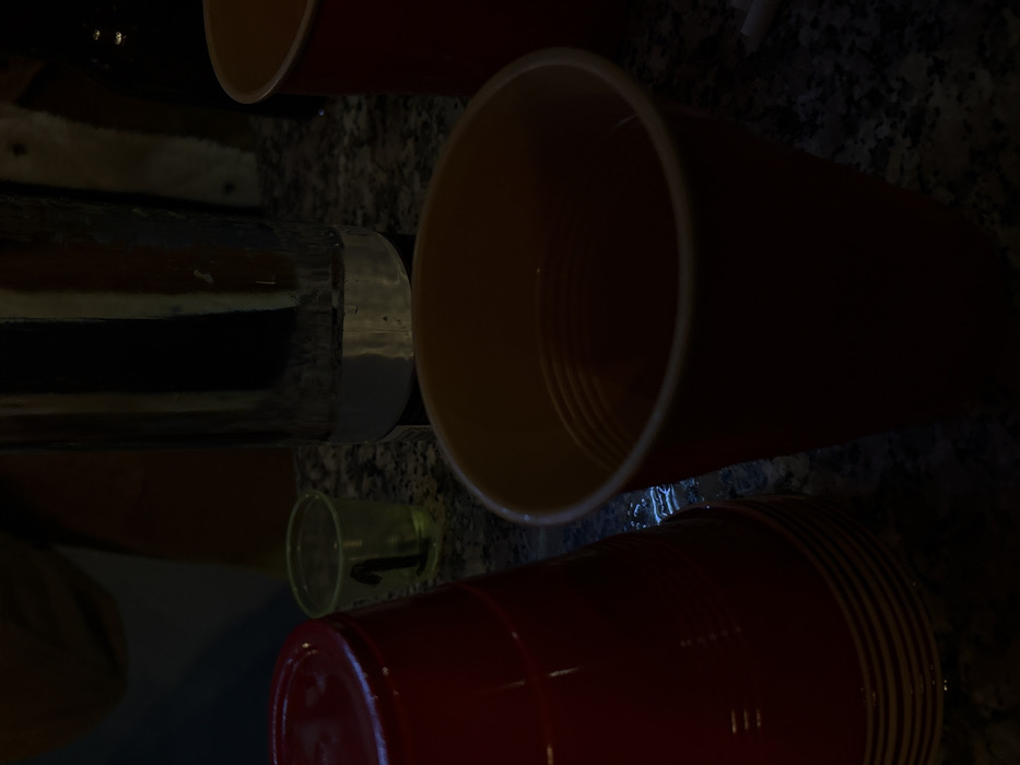 An array of items is present, including a stack of red plastic cups, a lone clear plastic cup bearing the number "1," and an empty orange plastic cup. Beside the cups, a tall, cylindrical object, possibly a glass or bottle, is visible. The setting appears to be a countertop with a granite-like finish, exhibiting dark grey and black speckles.
