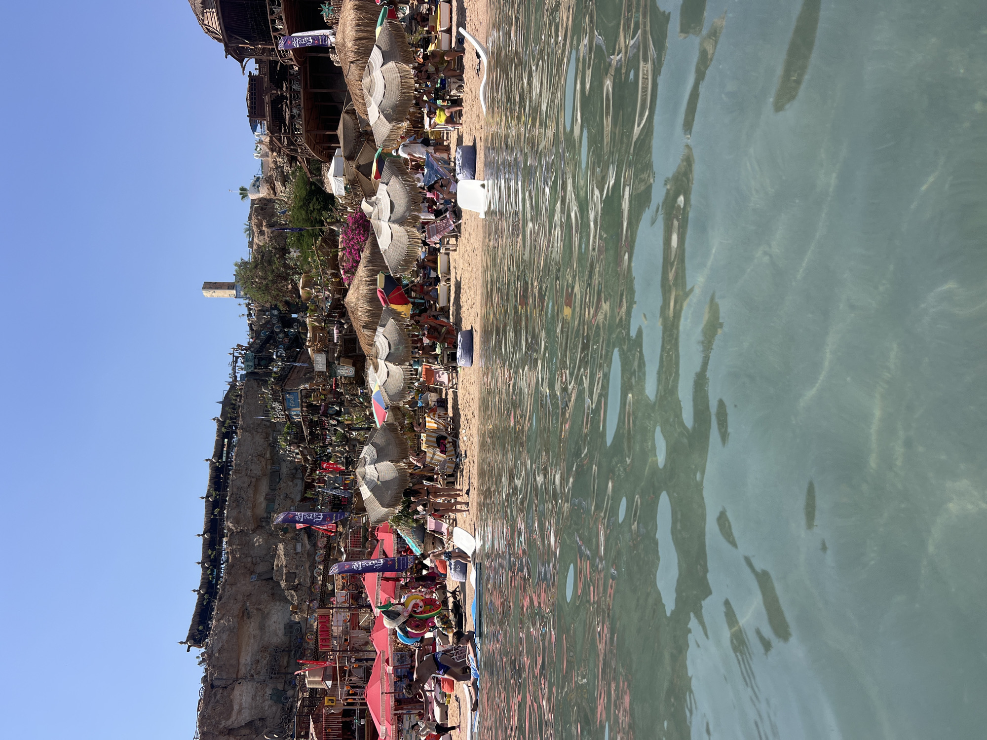 This is a photograph of a busy beach scene in Sharm El Sheikh, Egypt, likely located at the popular Naama Bay area. The image is taken from the water, looking towards the shore.
In the foreground, the clear, turquoise water allows visibility of the sandy seabed and several small fish swimming. The water's surface is gently rippling, reflecting the light.
The beach is crowded with people relaxing under numerous straw umbrellas and colorful awnings. Lounging chairs and beanbag-style seating are visible, indicating a recreational environment. People are depicted in various states of relaxation, some sitting, some lying down, and some appearing to be walking along the shore. There are children present, one lying on a flotation device in the water and another in a white swimming suit.
The background features a dramatic rocky cliff face with structures built into it, suggesting a resort or entertainment complex. These structures have a rustic, eclectic aesthetic, adorned with flags, lanterns, and decorative items. There are multiple levels of terraces and balconies with seating areas. The architecture appears to be a blend of traditional and modern styles, designed to integrate with the natural landscape.
Several tall, slender flags with writing on them are planted in the sand. Red and pink umbrellas are prominent among the beige straw umbrellas.
The overall atmosphere is lively and summery, with a clear blue sky overhead. The scene captures a popular tourist destination, characterized by its vibrant beach life, unique cliffside architecture, and clear waters.
