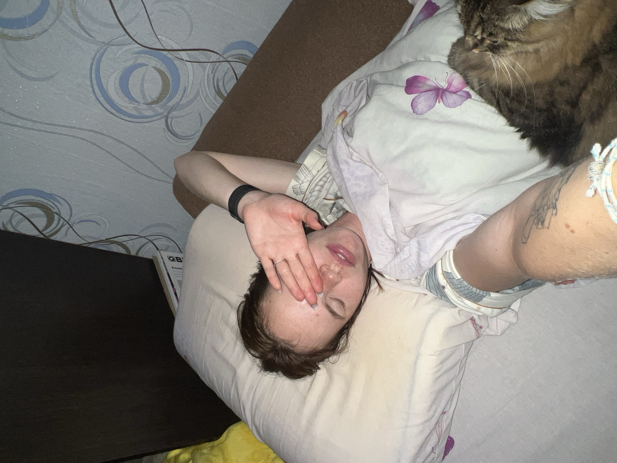 A young woman with brown hair is lying in bed, covering her eyes with her right hand. She is wearing a white shirt with a patterned design, and a blanket with a floral pattern is on top of her. A cat with tabby fur is also in the bed, partially visible in the lower right corner of the image. The background includes a dark headboard, wallpaper with a circular pattern, and some electrical wires. The scene appears to be indoors, likely in a bedroom, in the city of Chaikovsky, Russia.