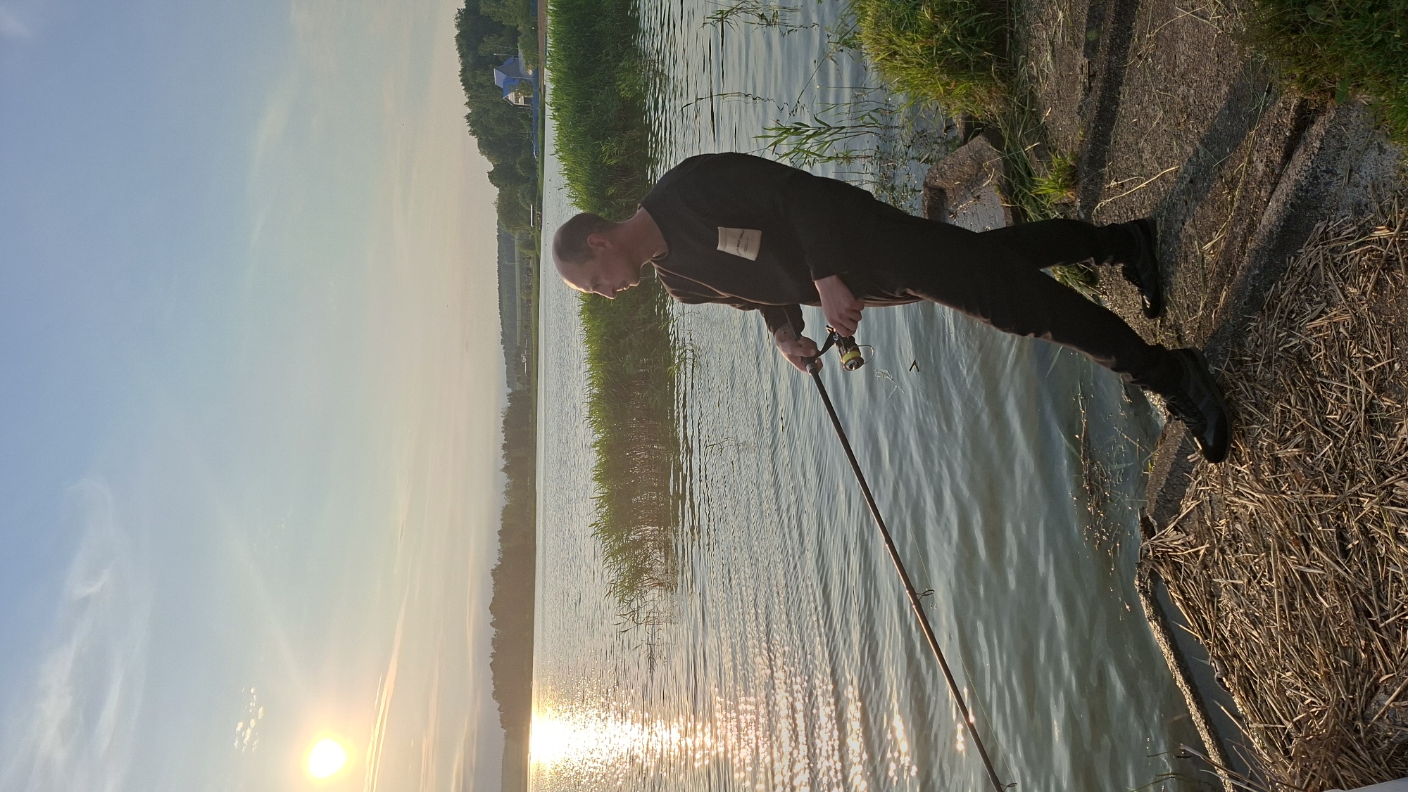 A man stands on a concrete embankment, fishing in a lake at sunset. The sun, low in the sky, casts a golden glow on the water, which is rippled and reflects the light. Tall reeds grow along the edge of the lake, and in the distance, a tree-lined shore is visible. The man is wearing dark clothing and is holding a fishing rod. He appears to be focused on the water. The scene is peaceful and serene, suggesting a moment of quiet contemplation. The location is likely a natural setting near the city of Smalyavichy, Belarus, given the presence of a lake and rural landscape.