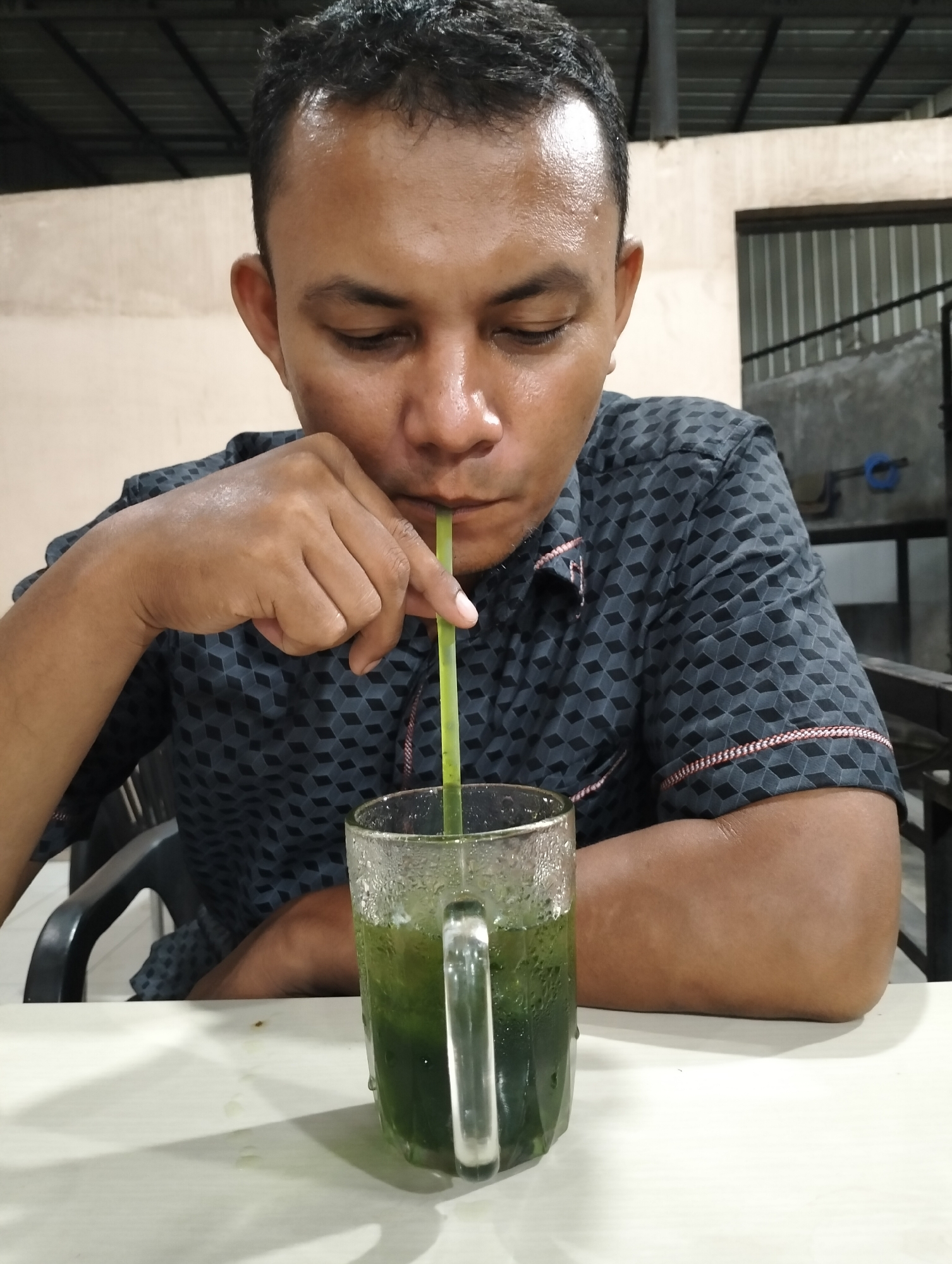 A medium shot depicts a dark-haired man drinking from a glass with a green-colored beverage and a green straw. The man is wearing a patterned black button-down shirt with red trim around the short sleeves. He is holding the straw with his right hand. The clear glass, covered in condensation, sits on a white table. Behind the man is a tan-colored wall. In the background, there is also a gray wall with a metal rack, and the metal rafters of a ceiling are visible overhead. The location is Lhokseumawe, Indonesia.