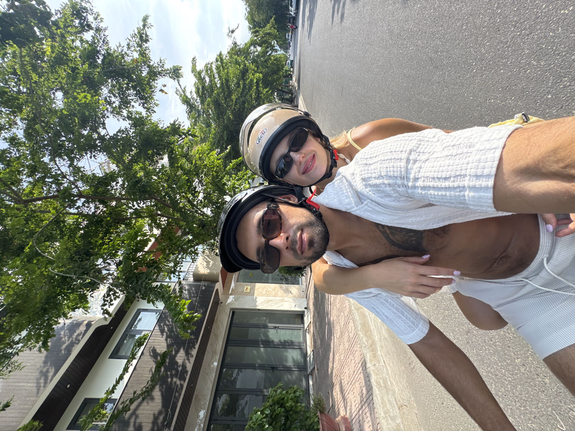 A couple is riding a scooter on a paved road in front of a modern, multi-story building in Nam Nha Trang Ward, Vietnam. The driver, a man wearing a black helmet and sunglasses, has his arm around the passenger, a woman also wearing a helmet and sunglasses. The woman is leaning on the driver's shoulder. Both are dressed casually in summer clothing. The building behind them has large glass doors and is partially obscured by lush green trees. The sky is bright and clear, suggesting a sunny day. The road is lined with more trees and other vehicles can be seen in the distance. The scene appears to be a typical urban environment in Vietnam.