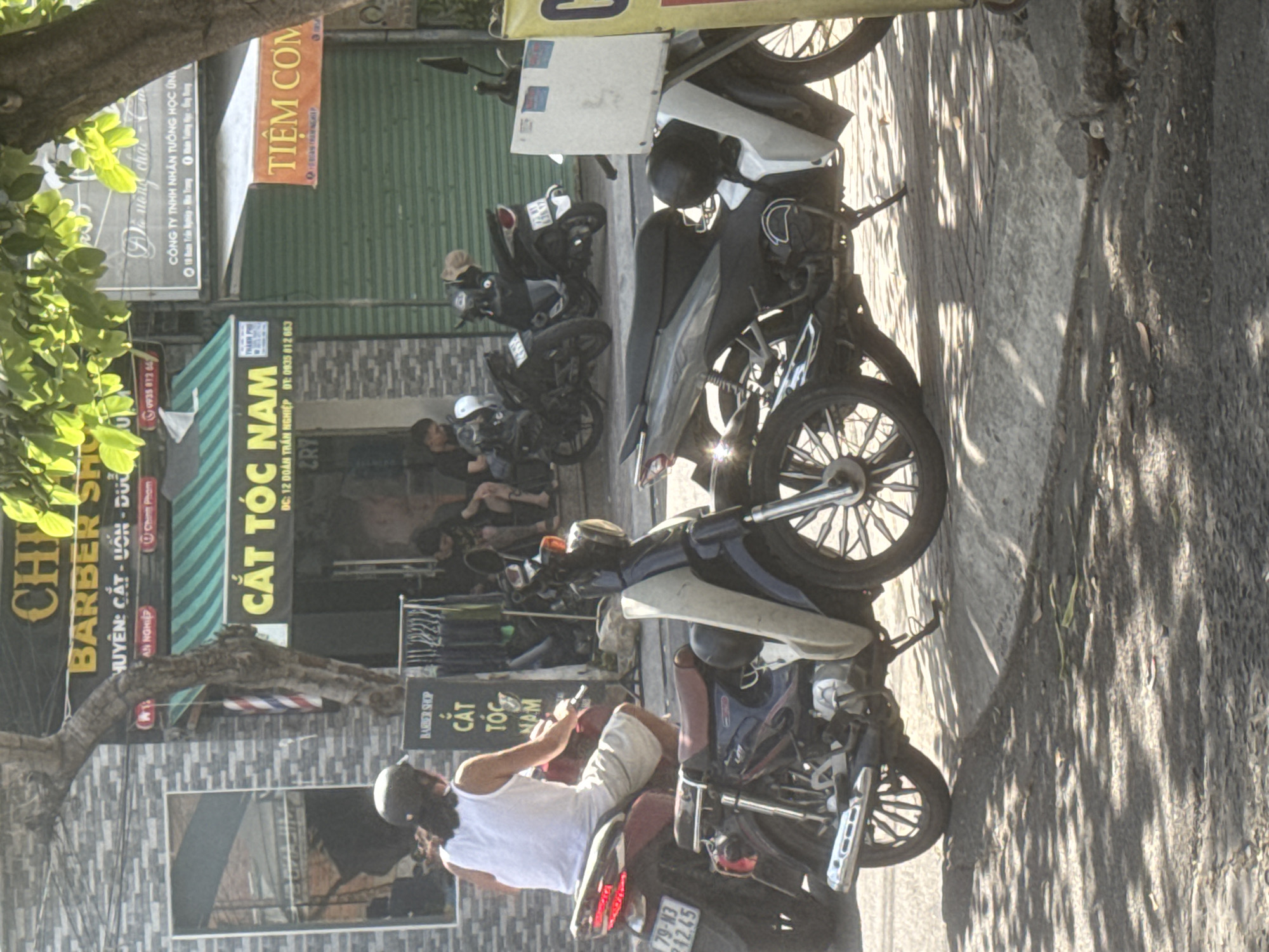 The image shows a street scene in Bac Nha Trang Ward, Vietnam. A person wearing a white tank top and light-colored shorts is riding a red motorcycle, facing away from the camera. The motorcycle has a license plate that reads "79-N3 742.45". In front of this motorcycle is a dark blue and white motorcycle with a person's legs visible on it. To the right of this is a gray motorcycle.
Behind the motorcycles, there are several shops. The shop on the left has a sign that says "CHILI BARBER SHOP" in large yellow letters and "CẮT TÓC NAM" (men's haircuts) below it. Another sign is partially visible, reading "TIỆM CƠM" (rice restaurant) in orange letters. The shops have green metal roll-up doors. There are also some people sitting outside the shops, one of whom appears to be a man with dark hair.
The ground is paved with concrete and some parts are shadowed by trees, creating a dappled light effect. The scene is outdoors on a sunny day.