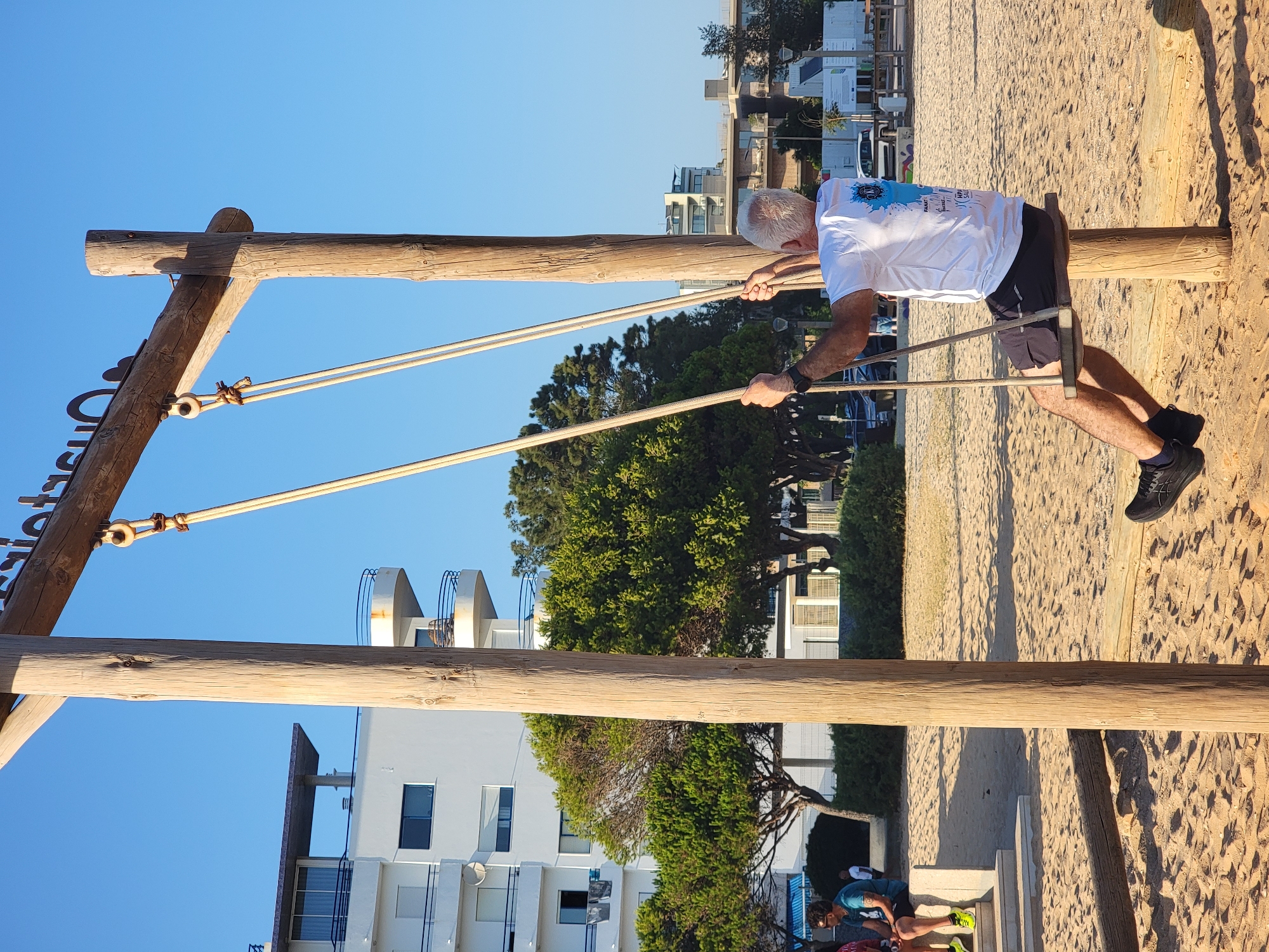 An elderly Caucasian man with gray hair is swinging on a wooden swing set at a beach in Quarteira, Portugal. He is wearing a white t-shirt with a blue graphic on the back and black shorts and black athletic shoes. The swing set is made of large wooden posts and a wooden beam, with ropes holding a wooden seat. The man is holding onto the ropes with both hands. The background shows a sandy beach, with some trees and modern buildings in the distance. The sky is clear and blue. The lighting suggests it is late afternoon. There are other people in the background, sitting and walking on the beach.