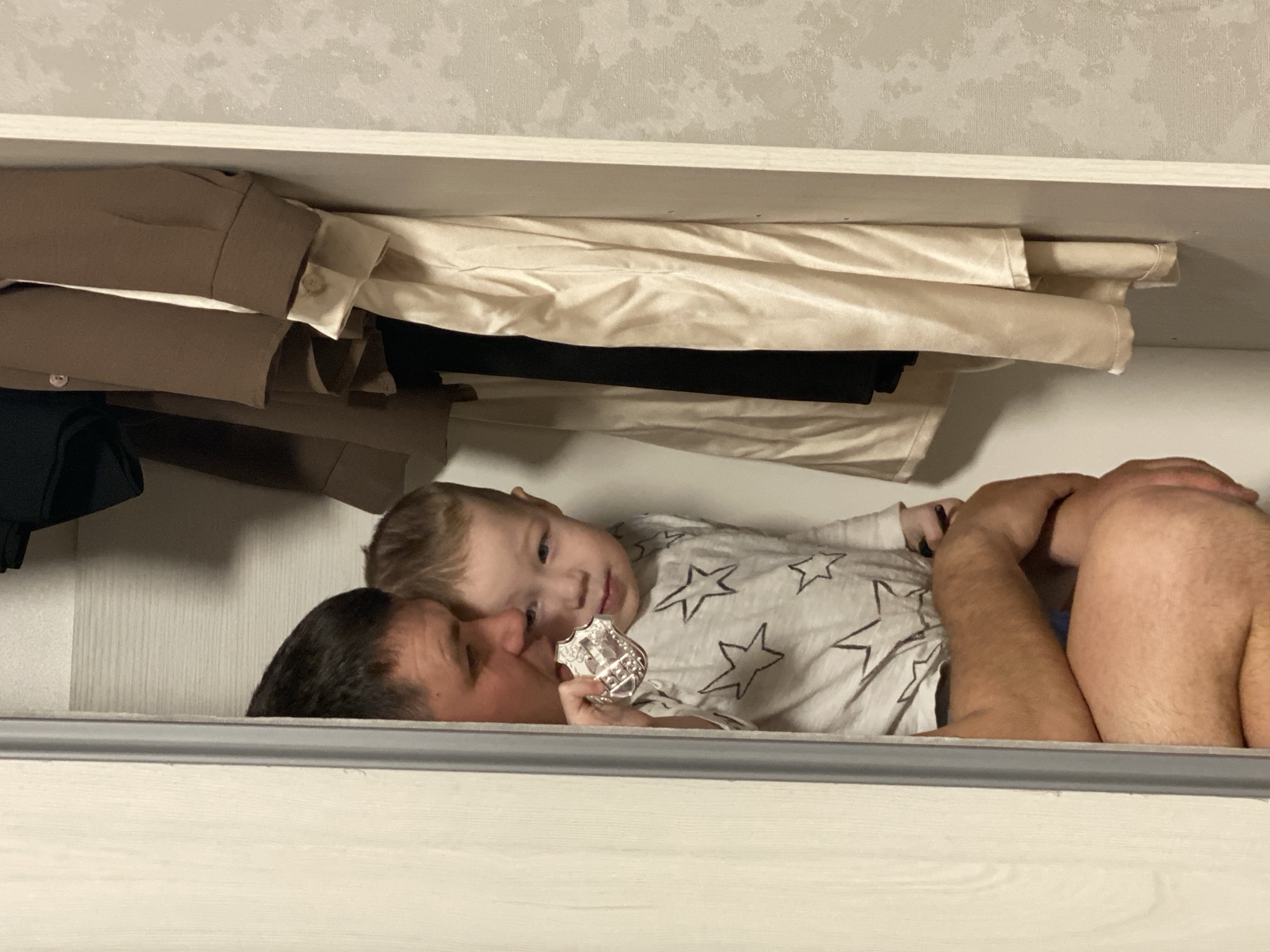 A toddler lies in an open closet with an adult male. The toddler, wearing a white long-sleeved shirt with grey stars, holds a silver-colored object in their left hand. The adult male lies beside the toddler. Clothes are stacked above them on the shelf. The scene's location in Sochi, Russia, cannot be determined from the image.
