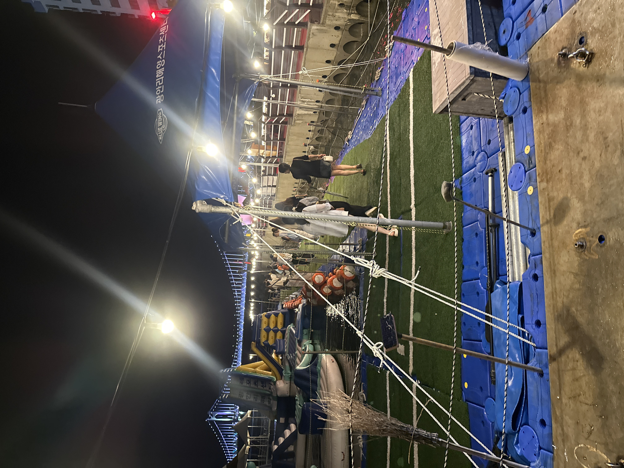 The image captures a nighttime scene at a water sports center in Busan, South Korea, likely situated near the Gwangalli Beach area, evidenced by the prominent Gwangandaegyo Bridge illuminated in the background. The foreground is dominated by a floating platform structure made of blue plastic modules covered with artificial turf, crisscrossed with white ropes. Several people are visible, some walking on the turf-covered platform, others near inflatable water structures or under a large blue tent. The tent has text that appears to be "BLUE WING" and "광안리해양스포츠센터" (Gwangalli Marine Sports Center). The water is dark, reflecting some of the lights. There are inflatable slides and structures floating on the water, suggesting a recreational area. The overall atmosphere is lively, with artificial lighting from the bridge, the tent, and string lights illuminating the scene.