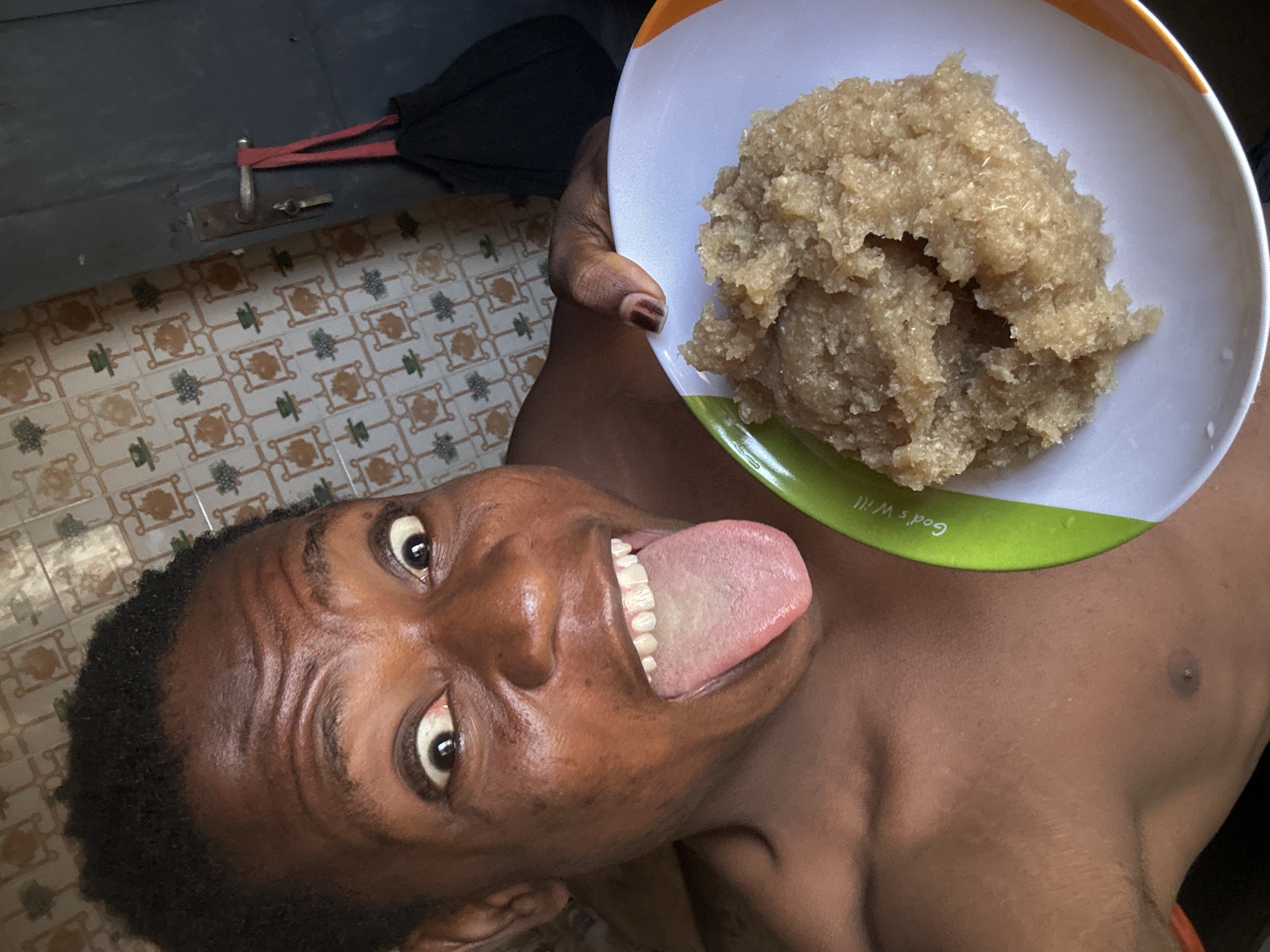 A shirtless man is holding a plate of food while standing in a room with tiled floors in an unknown location in the city of Unknown, Unknown. The man has dark hair, brown skin, and is sticking out his tongue while looking at the camera. He is holding a white plate with an orange rim and green base, on which there is a pile of a beige-colored, textured food. In the upper left corner of the image, there is a black bag or umbrella with red straps resting on a dark surface. The tiled floor is visible beneath the bag and features a pattern of brown and green geometric shapes on a light background.