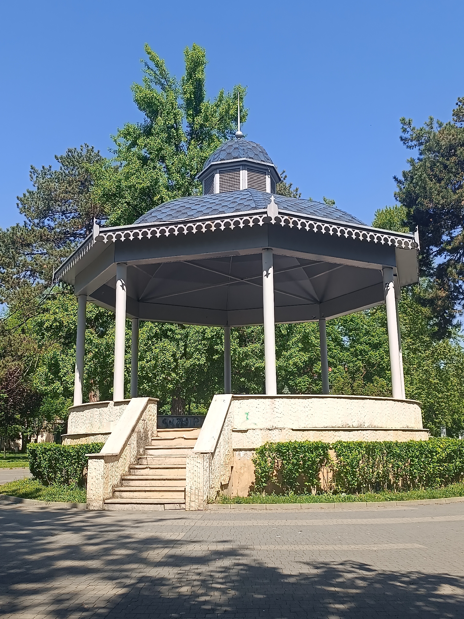 The image shows a gazebo in the city park of Pleven, Bulgaria. The gazebo has a stone base, columns, and a dark shingle roof with decorative trim. Steps lead up to the gazebo on two sides. The gazebo is surrounded by green trees and bushes. The ground is paved with gray brick, and shadows are visible on the brick. The sky is blue.
