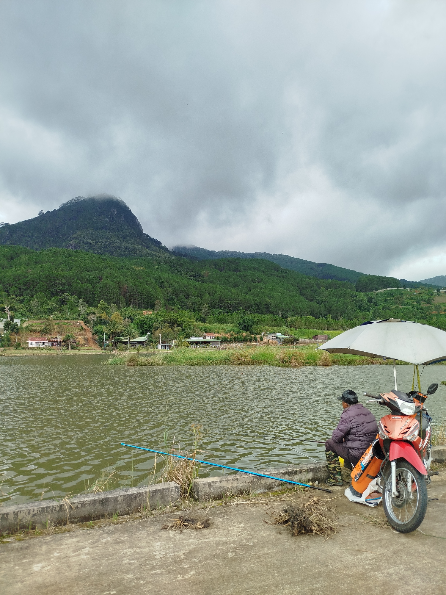 In the city of K'Long, Vietnam, on a cloudy day, a person is fishing by a lake. The person is seated on a concrete embankment next to a red and orange motorcycle. An open, light-colored umbrella is positioned behind the person and the motorcycle. A blue fishing rod extends into the greenish water. The background features a forested mountain peak partially obscured by clouds, with some buildings visible at the base of the mountain. The foreground consists of the concrete embankment, dry vegetation, and the lake's surface.