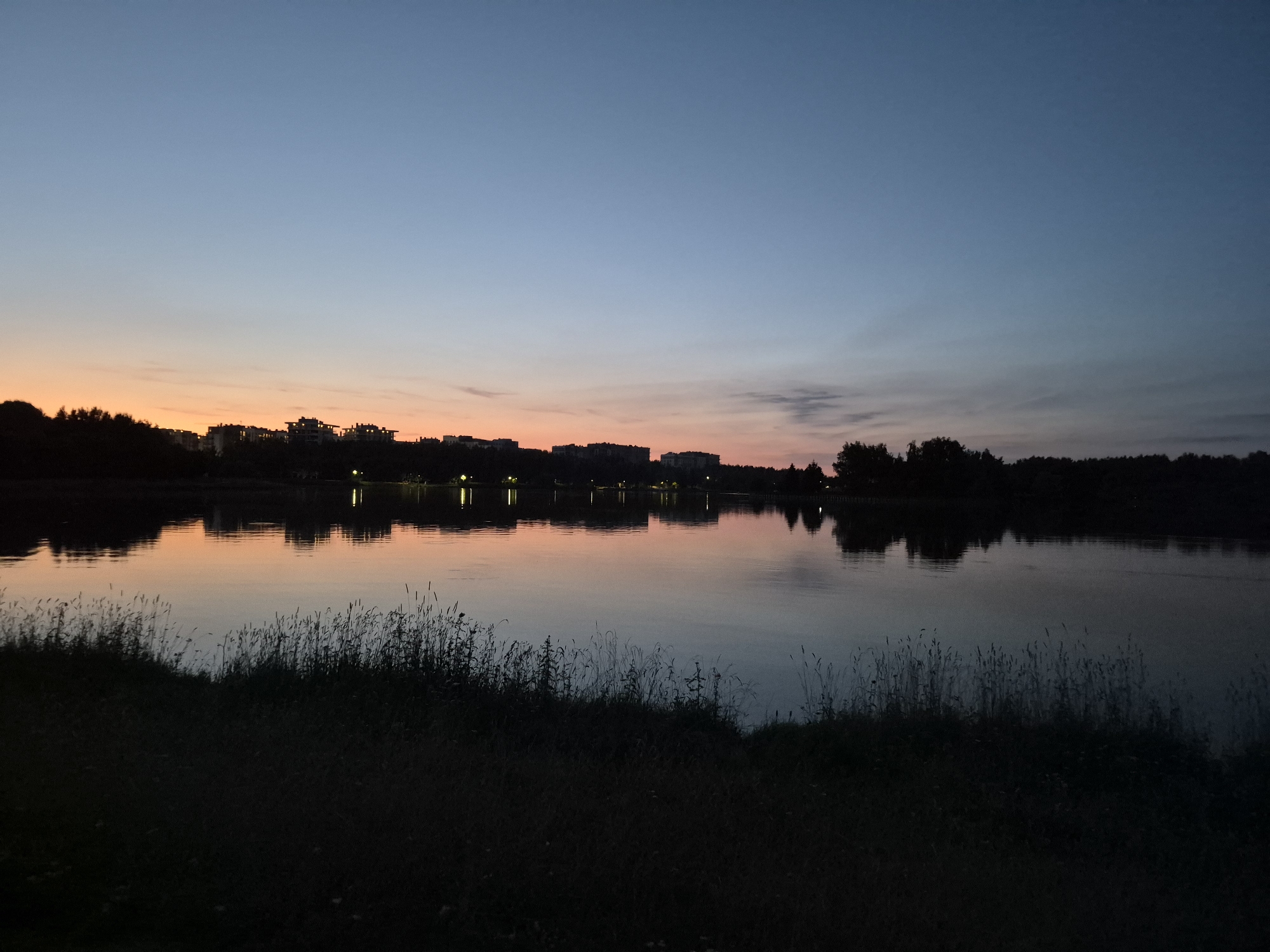 A wide-angle photograph captures a tranquil lake scene at dusk in Minsk, Belarus. The sky exhibits a gradient of colors, transitioning from a pale orange near the horizon to a deep blue overhead, with faint clouds adding texture. Across the lake, a silhouetted treeline marks the distant shore, punctuated by the faint outlines of buildings and scattered lights. These structures reflect on the calm surface of the water, creating shimmering distortions. The foreground is dominated by the dark silhouette of tall grass and vegetation lining the lake's edge, obscuring any definitive landmarks or identifying features of the specific location within Minsk. No people or discernible activities are present in the frame.