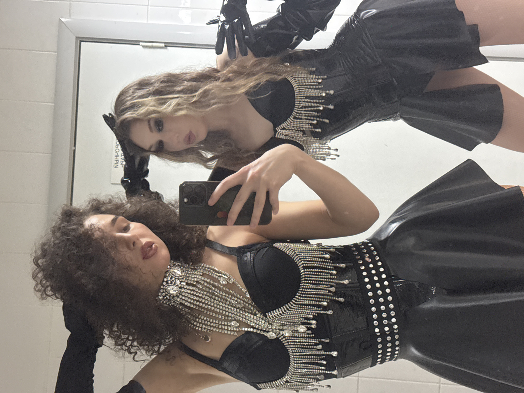 Two individuals are posing for a selfie in a room with white tiled walls and a reflective surface, possibly a mirror or a glass door. The person on the left has curly dark hair and is wearing a black corset with silver fringe detailing, a studded belt, and a sheer black skirt. They are also wearing long black gloves and a prominent silver necklace with crystals. The person on the right has wavy blonde hair and is wearing a similar black corset with fringe, a studded belt, and a short black skirt. They are also wearing long black gloves. Both individuals have their mouths slightly open as if posing. The background includes what appears to be a sign reflected in the surface, but the text is not legible. The lighting suggests an indoor setting, possibly a dressing room or bathroom. There is no indication of the location being Kazan, Russia.