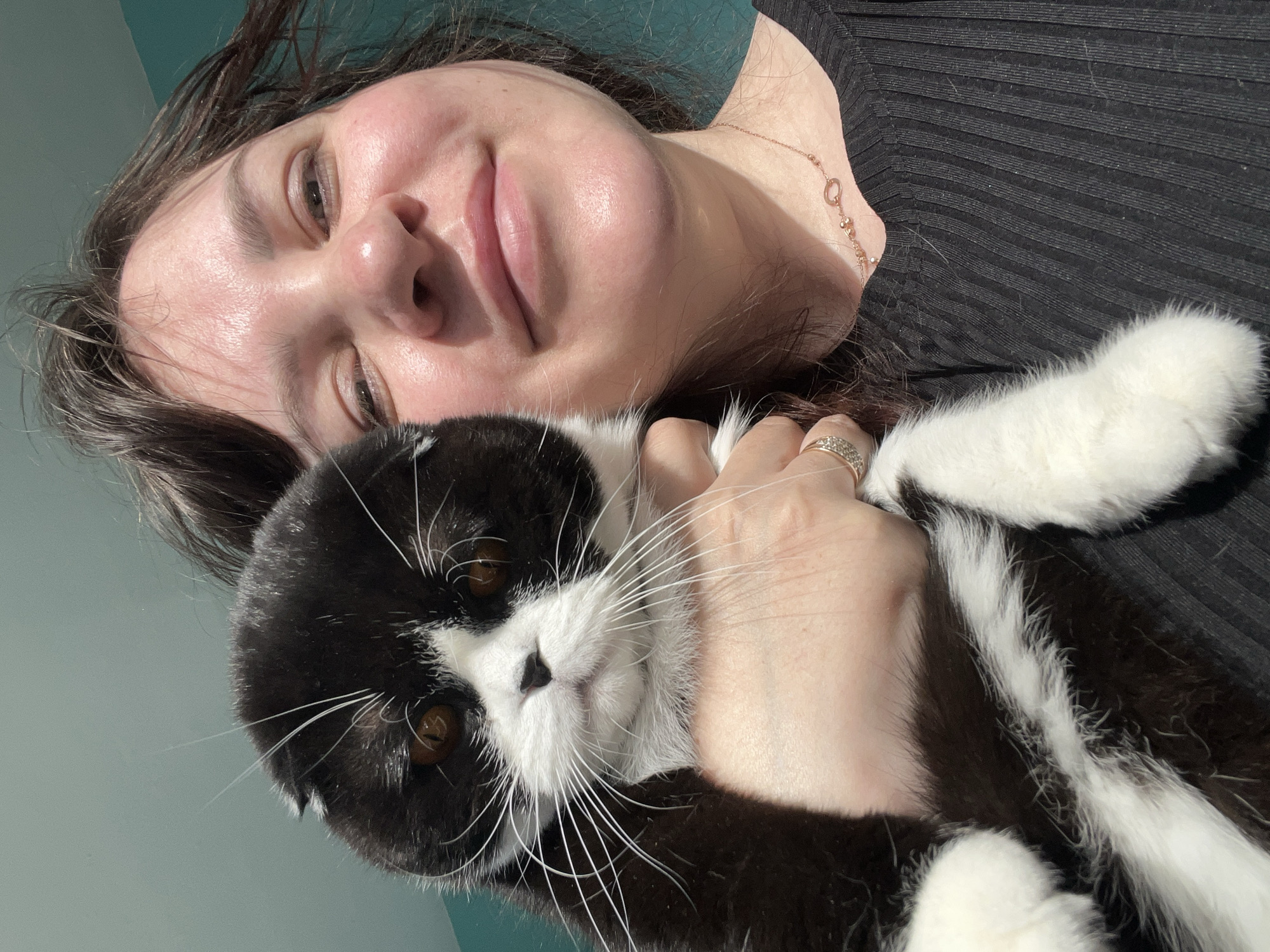 A selfie shows a light-skinned female holding a black and white cat. The woman has brown hair and is wearing a black top and a necklace. She is wearing a ring on her finger. The cat is lying on its side. No evidence to support the location of Perm, Russia is available from the image.