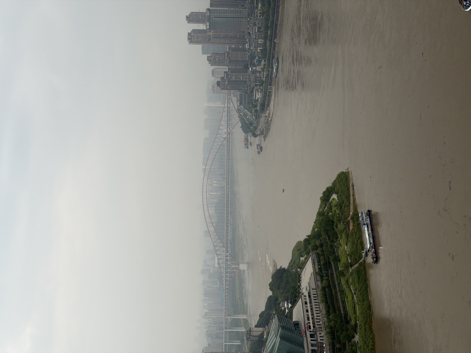 This is an aerial, wide-angle photograph of a cityscape on a cloudy day, likely taken from a high vantage point. The scene depicts a wide, muddy river flowing through a densely populated urban area. On the left side of the river, a green, sloped park area borders a large, modern building with a distinctive green, angular roof. Further into the distance on the left, a bridge with a red arch spans the river, connecting to a hazy cityscape of numerous high-rise buildings. On the right side of the river, another section of the city rises with a cluster of tall, modern buildings, one of which features prominent Chinese characters and the logo "ICBC". Several boats of varying sizes are visible on the river, with one larger, blue and white vessel docked near the green park area. The overall atmosphere is one of urban development situated within a substantial riverine environment, with limited visibility due to the overcast sky and atmospheric haze. The location is the Yuzhong District, China.