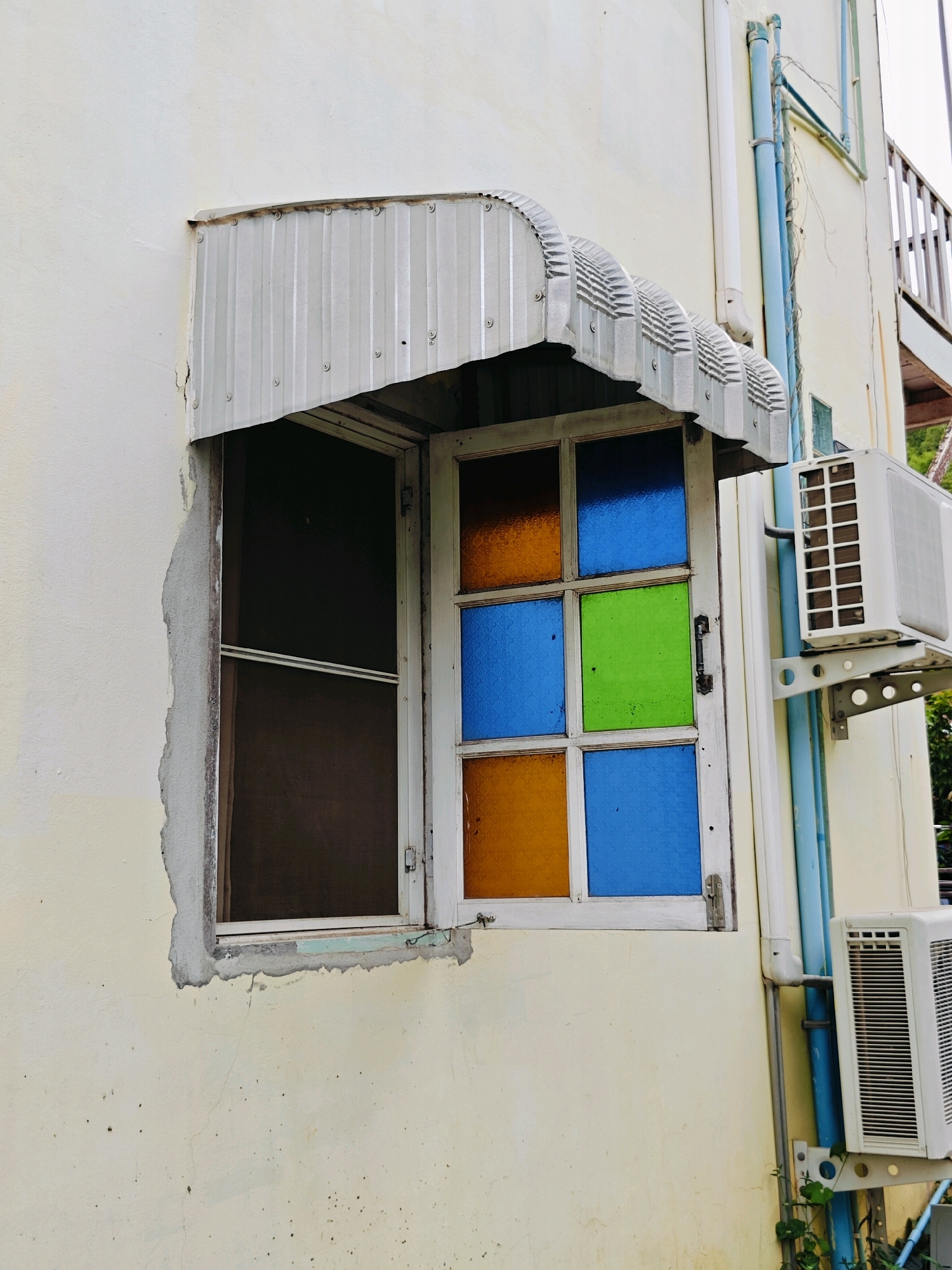 A close-up, eye-level, outdoor shot captures a detail of a building's exterior in Khao Yai, Thailand. The scene focuses on a cream-colored wall with a window assembly. Above the window, a corrugated metal awning with a scalloped edge protrudes. The window itself is divided into six panes of colored glass: two orange, two blue, and one green. The window frame appears to be made of white-painted wood, and a dark brown screen is visible behind the glass panes. To the right of the window, mounted on the wall, are two white air conditioning units, one larger and higher than the other. Blue and gray pipes run vertically alongside the units, attached to the building. The wall shows signs of wear, with some chipped paint and minor cracks. The overall impression is one of a functional, somewhat weathered, exterior detail.