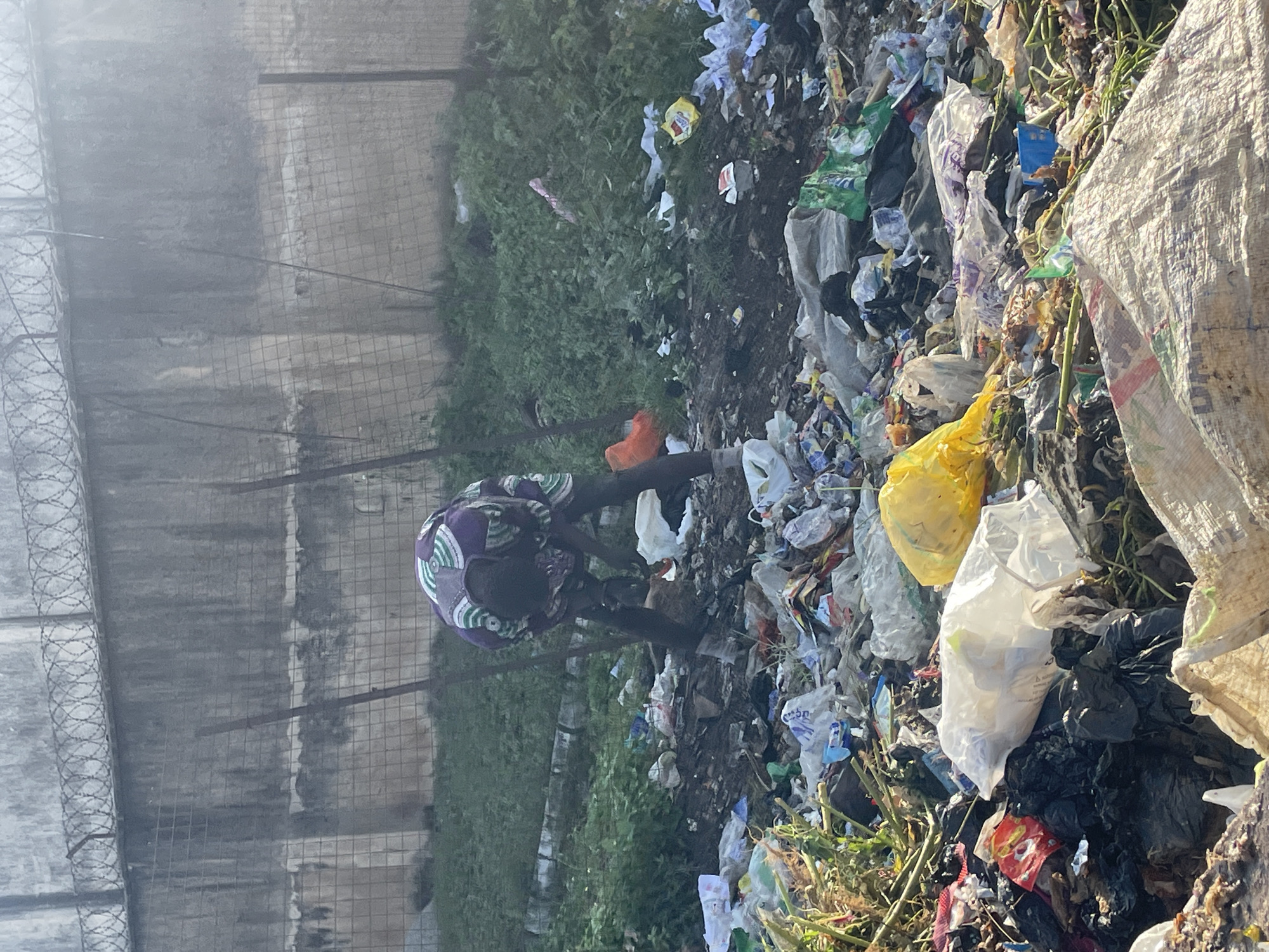 A person wearing a purple and white patterned shirt and dark pants is bending over in a large pile of trash. The trash consists of various plastic bags, containers, and other debris. In the background, there is a concrete wall topped with barbed wire, and some sparse vegetation. The scene appears to be an outdoor dump site in Barakin Lerawi, Nigeria.