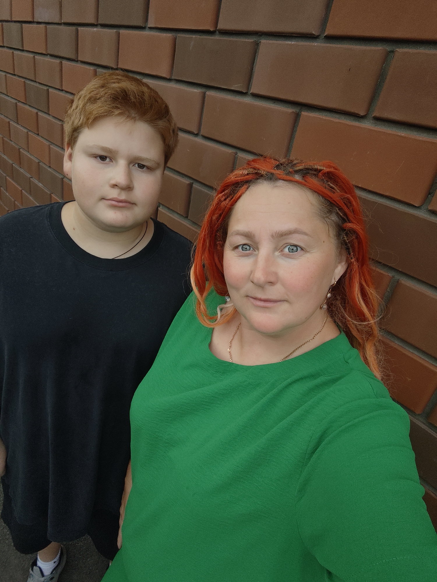 The image shows a medium shot of two people, a younger person with red hair on the left, and an older person with bright orange hair on the right, standing in front of a brick wall. The younger person is wearing a black t-shirt and the older person is wearing a green top. Both are looking at the camera. The background is a brick wall with a textured, reddish-brown pattern. There are no other discernible features of the environment or specific location.