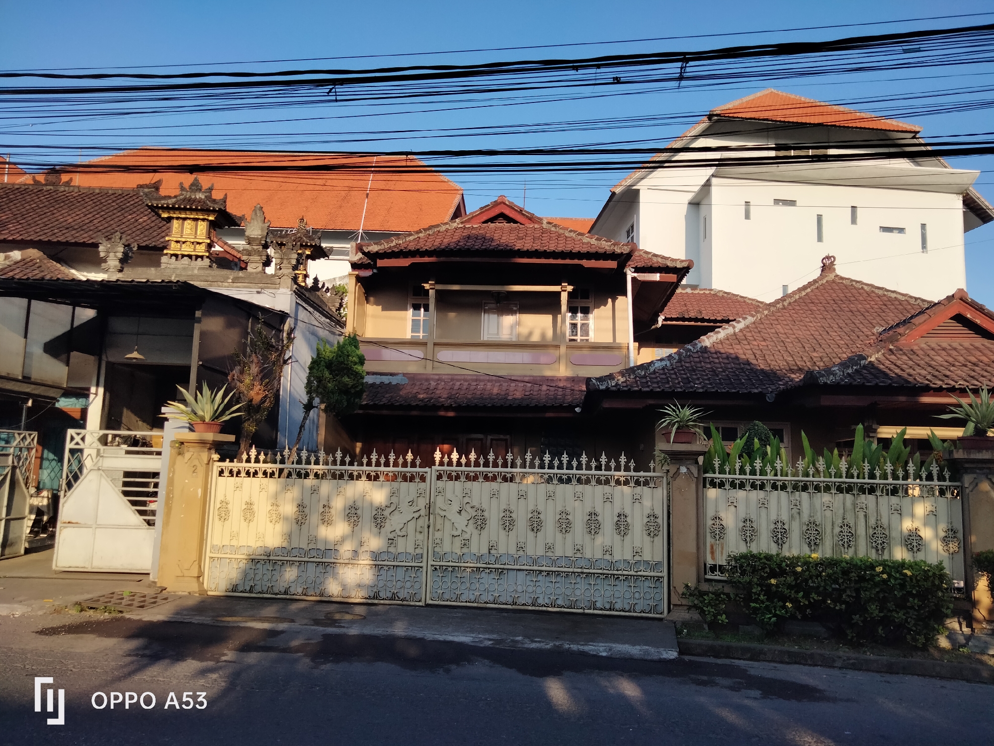 The image shows a street scene in Denpasar, Indonesia, with a series of residential buildings and their fences. The architecture appears to be a mix of traditional Balinese and modern styles, with terracotta tile roofs dominating the skyline. The foreground features a paved road with shadows cast by overhead power lines and the buildings.
A prominent feature in the image is an ornate, cream-colored metal gate, intricately designed with decorative patterns. To the left of this gate, there is another, simpler white metal gate, and a portion of a white building can be seen behind it. The gate is flanked by fences, also made of metal, adorned with similar decorative elements.
Several potted plants and bushes are placed along the fence and near the gate, adding a touch of greenery to the scene. Behind the gates and fences, the buildings exhibit varying architectural styles. Some have multi-story structures with balconies and windows, while others have more elaborate roof designs featuring intricate carvings and finials, typical of Balinese temples or ornate homes. A larger, modern white building with a simple facade and small, rectangular windows stands out amongst the other structures.
The sky is clear and blue, indicating a sunny day. The lighting suggests it is either early morning or late afternoon, given the long shadows. The overall impression is that of a residential area in a tropical climate, characterized by distinct architectural elements and dense overhead wiring. The "OPPO A53" watermark in the bottom left corner indicates the image was captured with this device.
