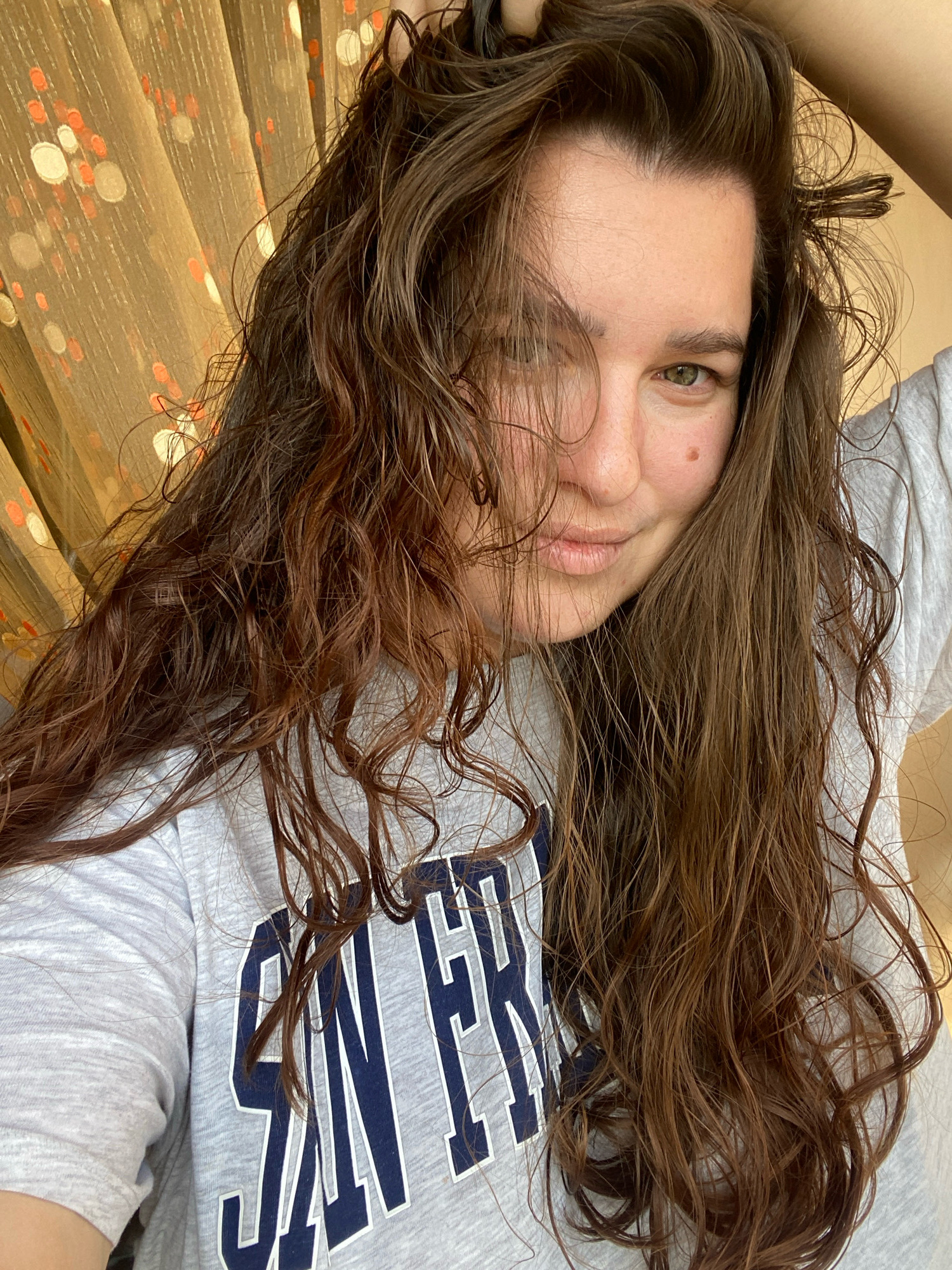 The image is a selfie taken by a young woman with long, dark brown, wavy hair. She is wearing a grey t-shirt with blue lettering that reads "SAN FR" on the chest. Her left hand is raised, touching her hair. Her right arm is bent, with her hand resting on her shoulder. The background is a patterned curtain with circular designs in orange and white. There are no visible landmarks or cues indicating a specific location in the city of Perm, Russia. The lighting suggests it might be daytime, possibly near a window due to the curtain.