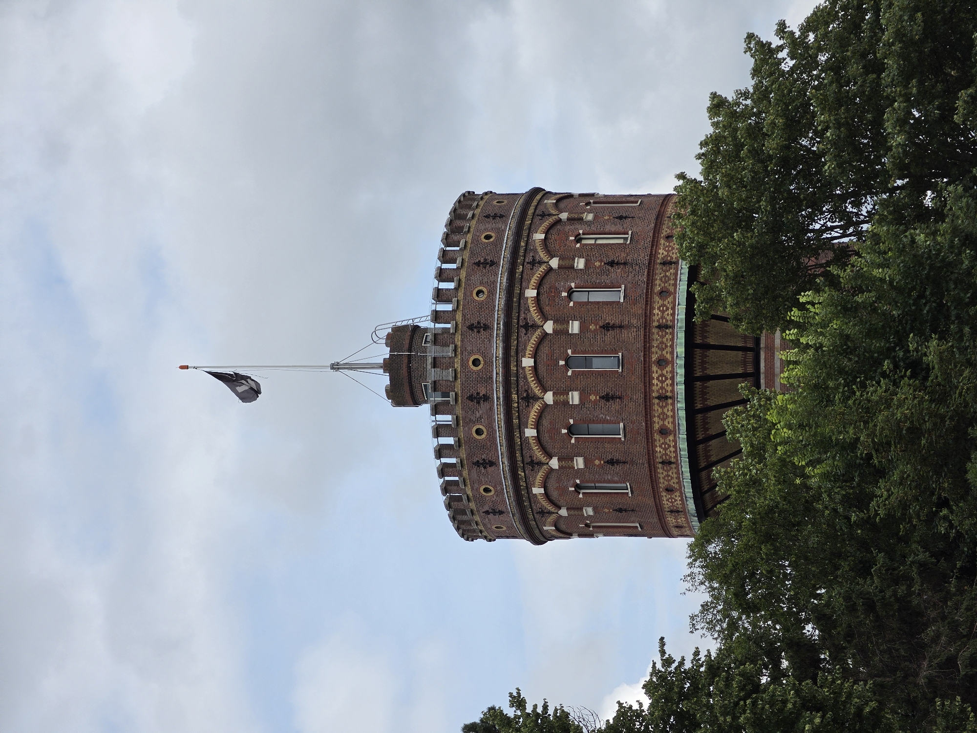 The image shows the top portion of a cylindrical brick water tower in Delft, Netherlands, partially obscured by lush green trees. The tower has a castellated parapet at the top, with small, dark windows and decorative brickwork, including arched window frames and circular accents. A flagpole with a black and white flag, possibly a pirate flag, stands at the very apex of the tower. The sky above is overcast with white and grey clouds. The trees in the foreground are dense and varied in color, suggesting a summer or early autumn setting.