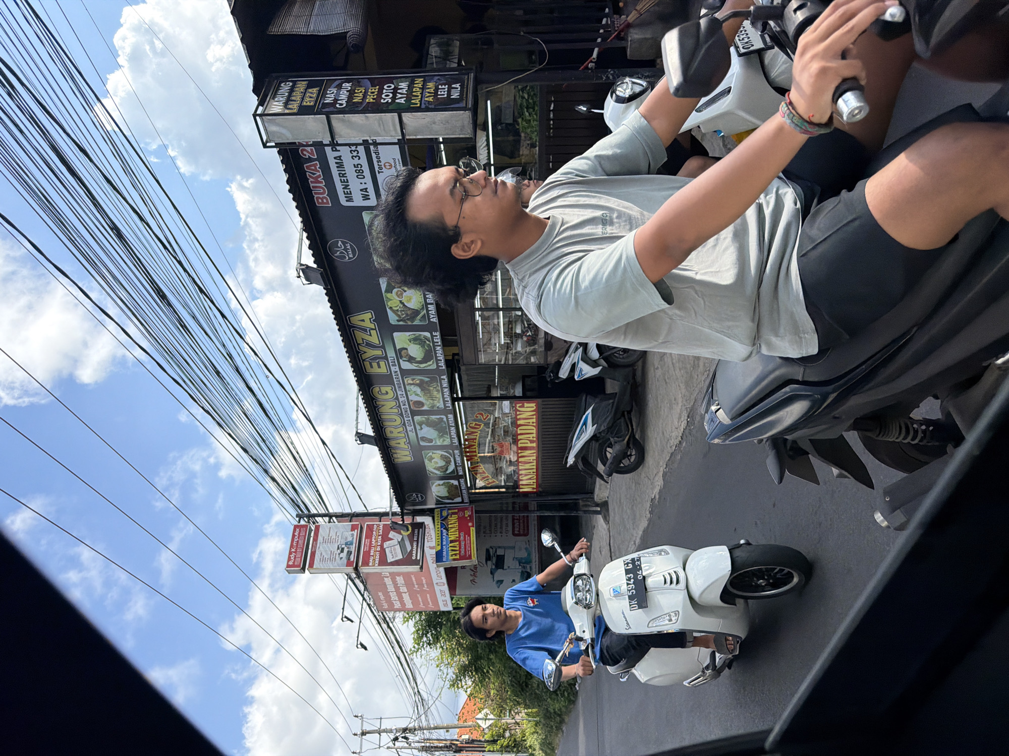 This daytime image, taken in Kuta Selatan, Indonesia, captures a street scene featuring two men on scooters. The primary subject, positioned in the foreground, is a man with dark curly hair and glasses, wearing a light grey t-shirt and black shorts. He is riding a white and grey scooter, with a partially visible license plate showing "DK 5399". His hands are on the handlebars, and he has colorful bracelets on his right wrist. Behind him, another man in a blue t-shirt rides a white Vespa-style scooter with the license plate "DK 5943 CX 04-27".
The setting is a bustling street lined with commercial buildings. The sky above is bright blue with scattered white clouds, and numerous overhead power lines crisscross the frame. Shopfronts are visible in the background, displaying various signs. One prominent sign belongs to "WARUNG EYZA", advertising Indonesian dishes like "NASI CAMPUR", "Nasi Pecel", "SOTO", "AYAM", "LALAPAN AYAM", "LALAPAN LELE", and "LALAPAN NILA", along with "MASAKAN PADANG". This warung also states it's "BUKA 24" (open 24 hours), accepts orders ("MENERIMA P"), and has a WhatsApp number. A "HALAL" logo is also visible. Another business, "Rudy Komputer", advertises computer services, laptop/HP sales on credit, and lists brands such as Acer, Lenovo, and Toshiba. The street appears to be busy with several other parked motorbikes/scooters. The overall impression is a typical day on an Indonesian street, with daily commute and local businesses in operation.
