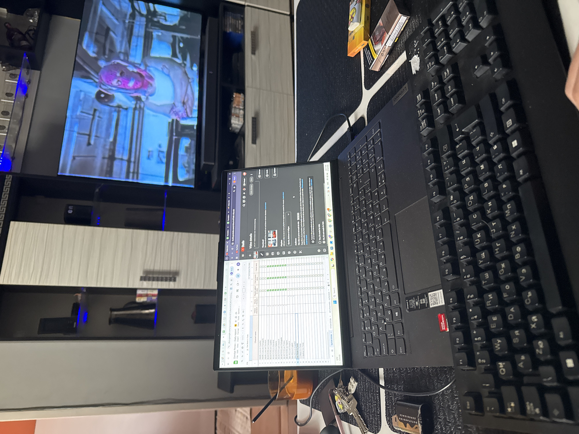 The image appears to show a desk setup, likely in an indoor environment. In the background is a TV screen displaying an image of a person with pink hair.
The main focus is on a laptop with an open spreadsheet on its screen, which is probably Google Sheets. A keyboard is also present, partially obscuring the laptop. Some personal items like keys, a glass with a straw, and cigarette packets are present on the desk. The spreadsheet appears to be a daily report, likely related to YouTube channels and video details.
It is not possible to determine the location in the city of Sofia, Bulgaria from the image.