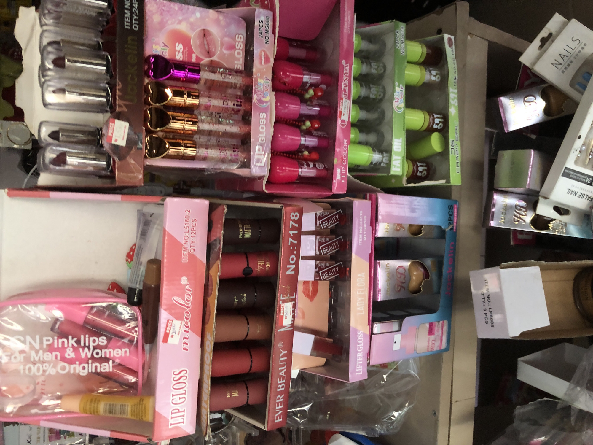 A close-up shot shows a cluttered shelf of cosmetics, presumably in Ado, Nigeria. The products include lip gloss, lipstick, and false nails. Brands visible are "Micolor," "Ever Beauty," "Lady Flora," "Miss Lovely," and "Jockelin." A pink cosmetic bag labeled "SN Pink Lips for Men & Women" is also present.