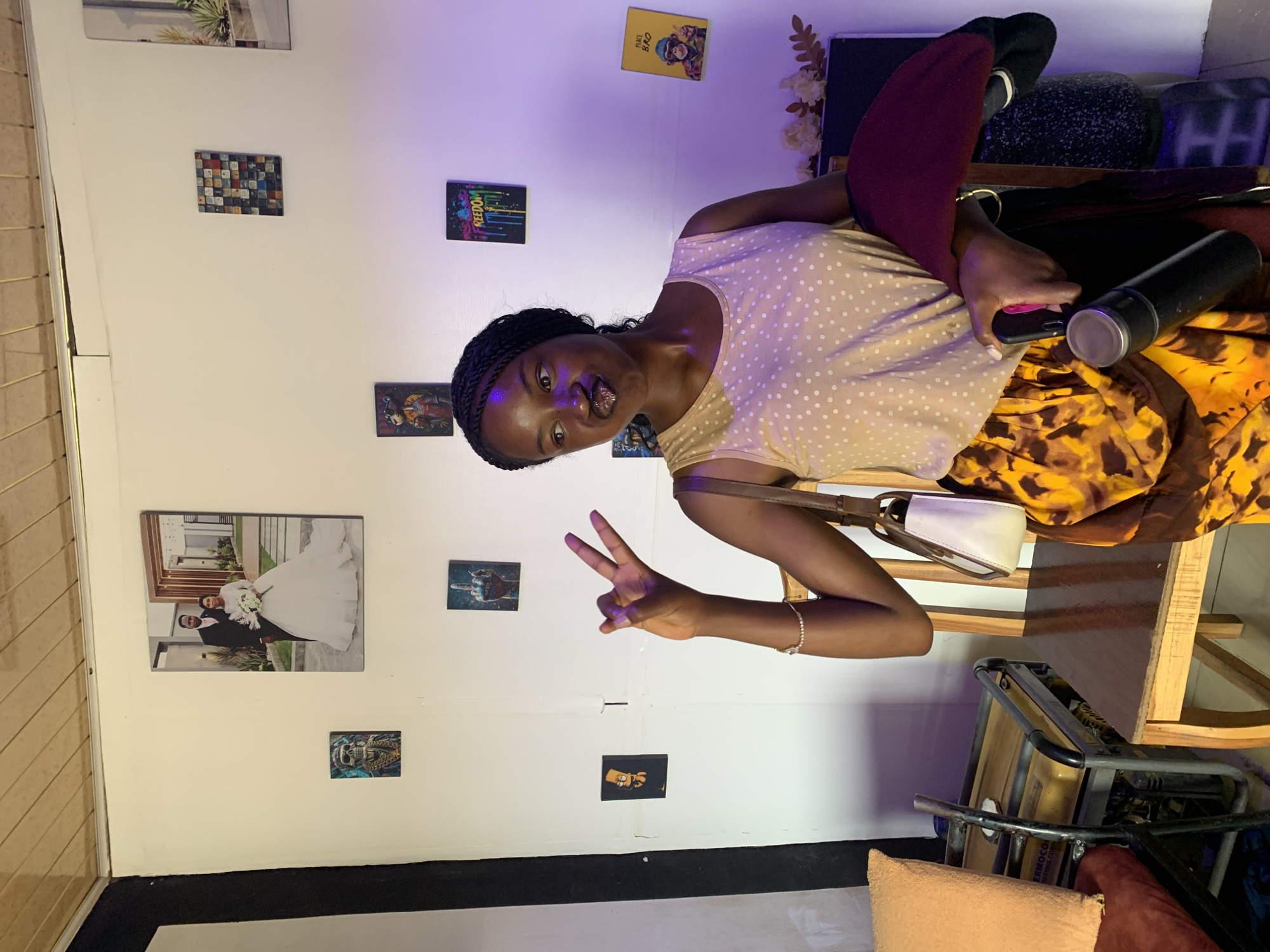 A young woman with braided hair is sitting indoors against a white wall decorated with various framed images. She's wearing a beige and white polka-dotted top and an orange and brown patterned skirt, with maroon leggings visible on her legs. She is holding a smartphone in her right hand, along with a silver thermos. She's holding up two fingers in a peace sign with her left hand. A small white purse with a brown strap is slung over her shoulder. The environment includes an assortment of items like a chair, pillows, and what looks like a portable generator in the background. Although the prompt requests the location in Jos, Nigeria, there is no information in the image to confirm this. Therefore, it is not possible to state whether the photograph was taken in Jos, Nigeria.