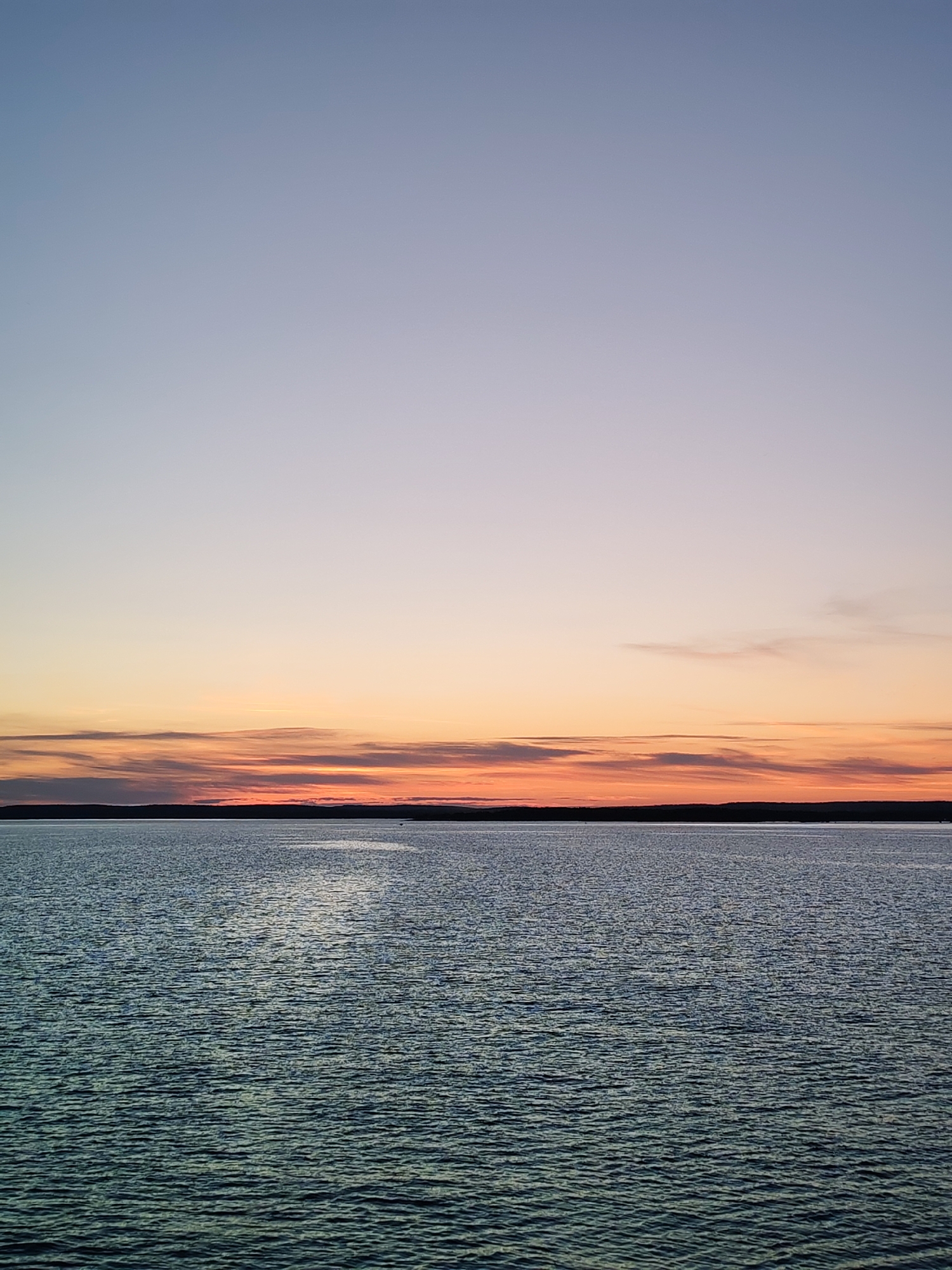 A serene seascape at dusk is depicted, with the sky transitioning through soft pastel hues of pale blue and light orange. A thin band of dark land, possibly a distant shore or island, divides the sky from the tranquil body of water. The water's surface is gently rippled, reflecting the fading light of the sunset, with a subtle sheen visible in places. The scene evokes a sense of peacefulness and natural beauty. This location is in Shal'skii, Russia.