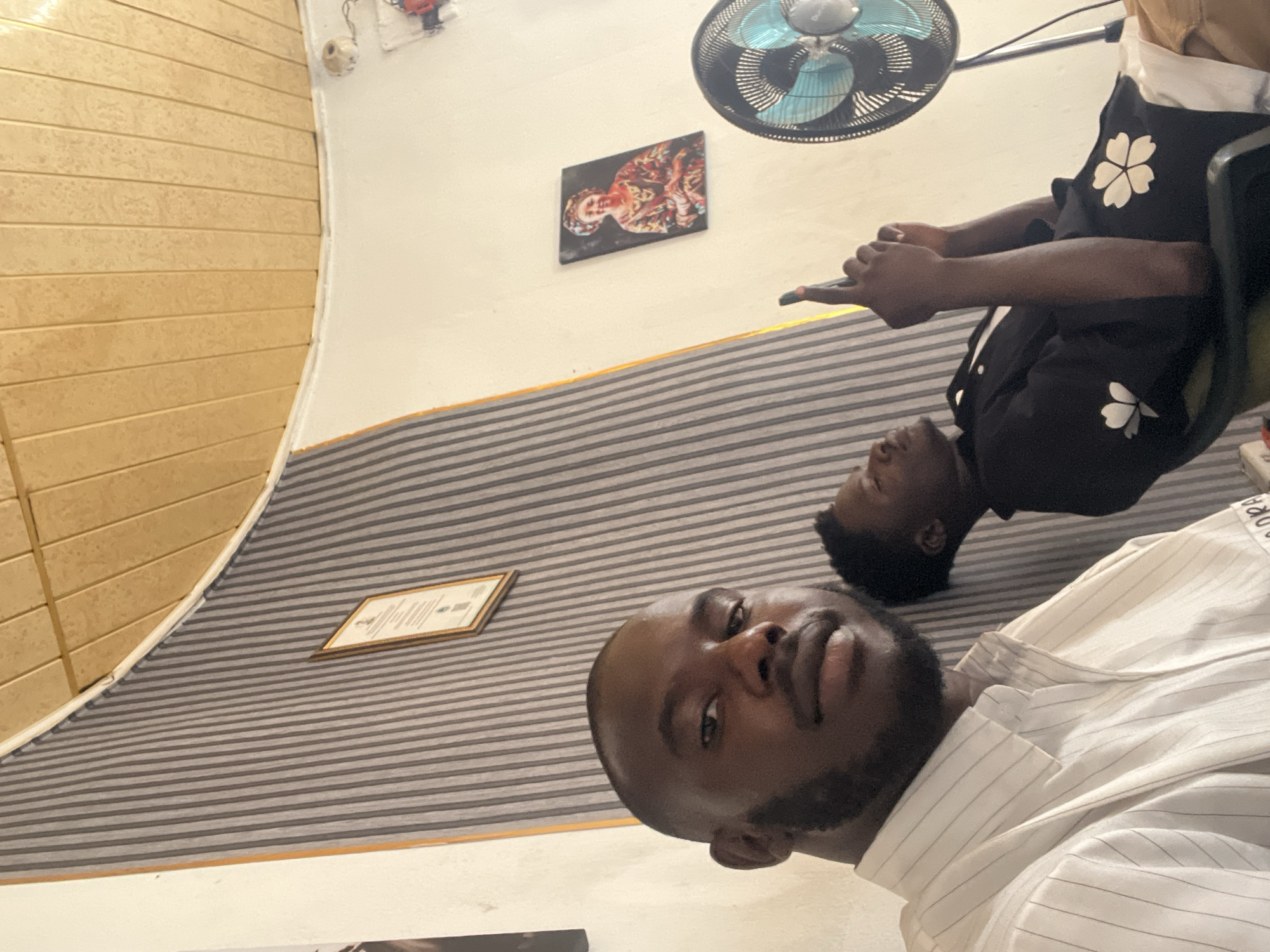 Two dark-skinned men are in a room. One man in the foreground is wearing a white striped shirt. The other man, in the background, is wearing a black shirt with white flower designs, and is holding a phone. There's a fan hanging above the men. The background is a room with a wall and wood paneling. A picture and certificate are displayed on the wall. The location is likely Gura Top, Nigeria.