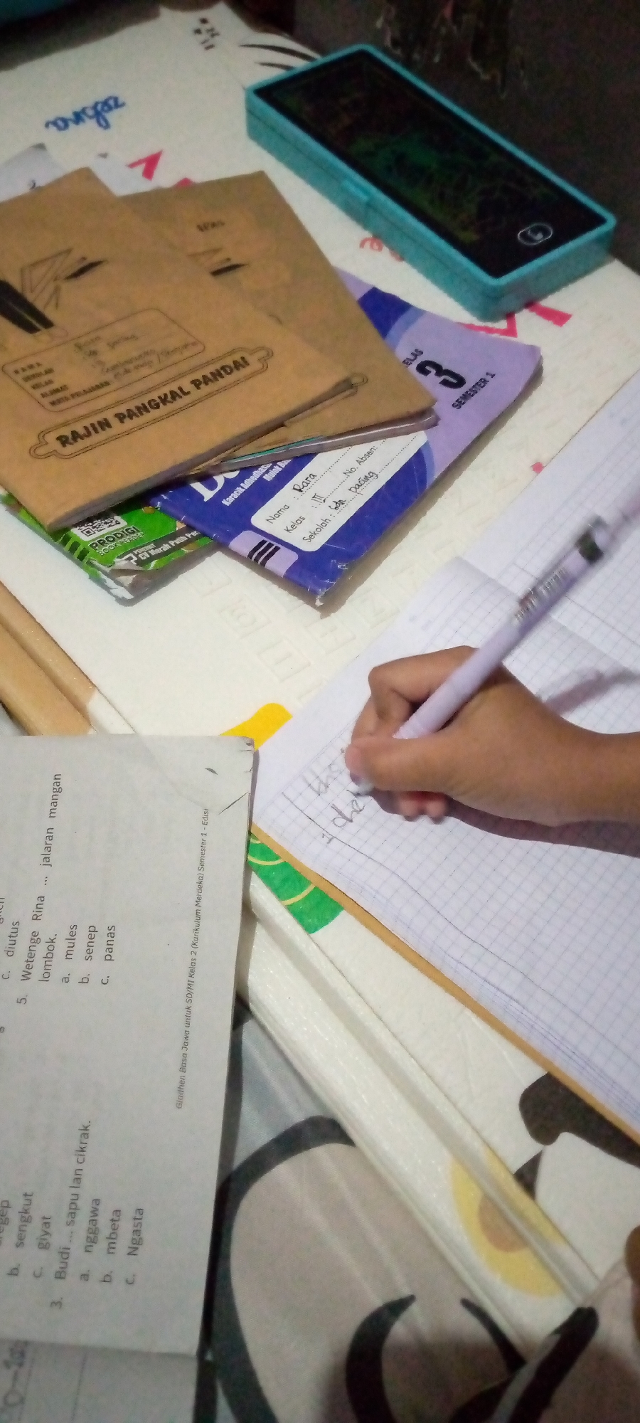 This image captures a close-up of a person's hand holding a light purple pen, writing in a grid-lined notebook. The notebook is open and placed on top of a stack of other books and papers. To the left of the notebook, a portion of a worksheet is visible, displaying multiple-choice questions in Indonesian. The questions include options such as 'diutus', 'sentrum', 'lombok', 'mules', 'senep', 'panas', 'sapu lan cikrak', 'nggawa', 'mbela', and 'Ngasta'. One of the books in the stack is brown with the text "RAJIN PANGKAL PANDAI" visible. Another book, purple and white, shows "SEMESTER 1" and the number "3". A turquoise electronic device, possibly a pencil case or a small tablet, is situated at the top right of the frame. The scene appears to be set on a patterned surface, suggesting a desk or table. The lighting is dim, and the overall image has a slightly blurred quality, with a focus on the writing action. The presence of Indonesian text on the documents indicates the location is likely in Indonesia. The provided context mentions "Sumberwono, Indonesia," which is a known location.