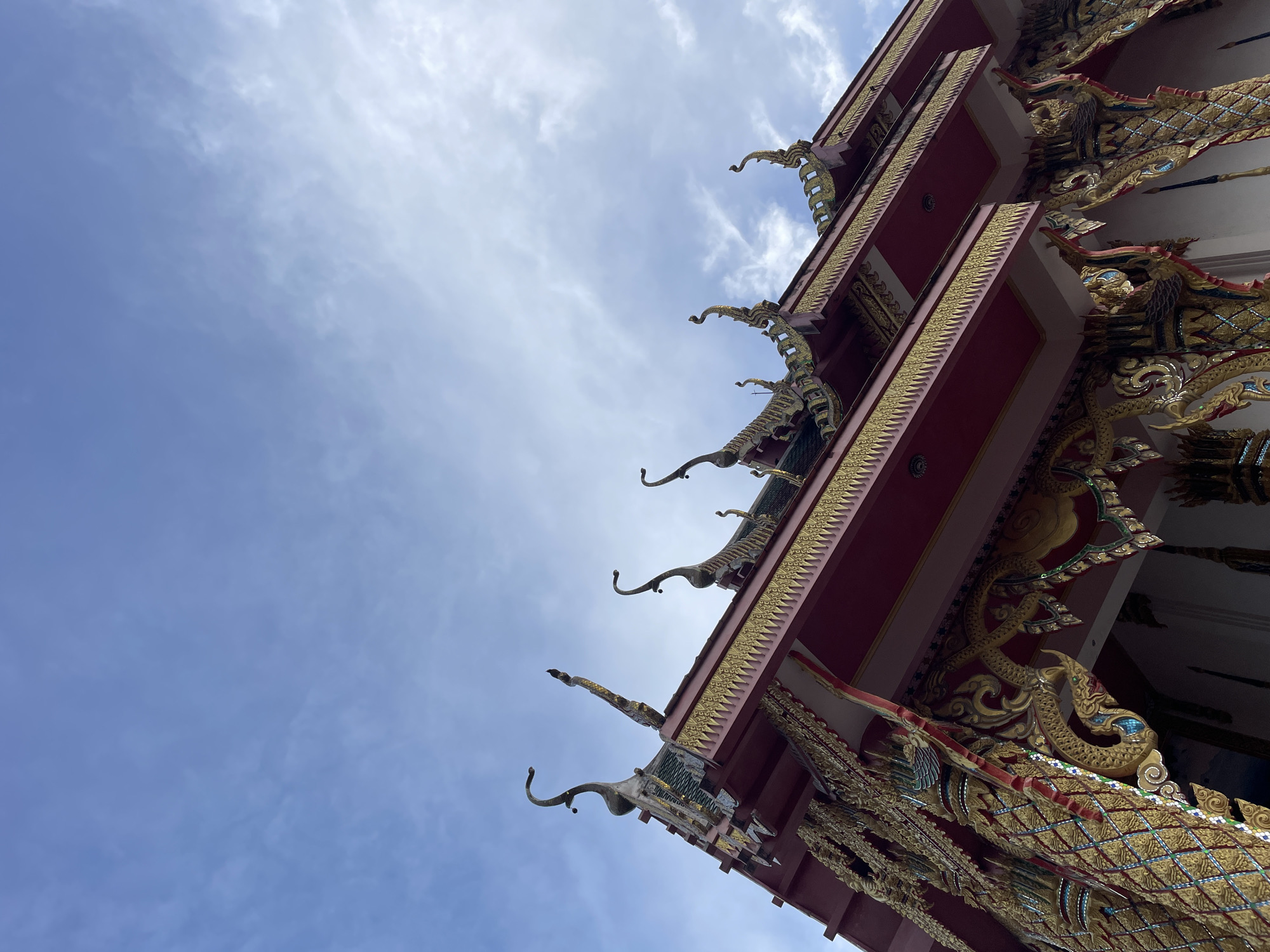 An upward angle shot captures the ornate roofline of a Thai temple against a pale blue, cloudy sky. The temple features a deep red underside to its eaves, adorned with intricate golden trim. The roof itself is decorated with mythical serpent-like figures with gold detailing, and stylized finials. Beneath the eaves, elaborate golden carvings depict dragon-like creatures with turquoise accents. The image was taken in Pattaya City, Thailand.