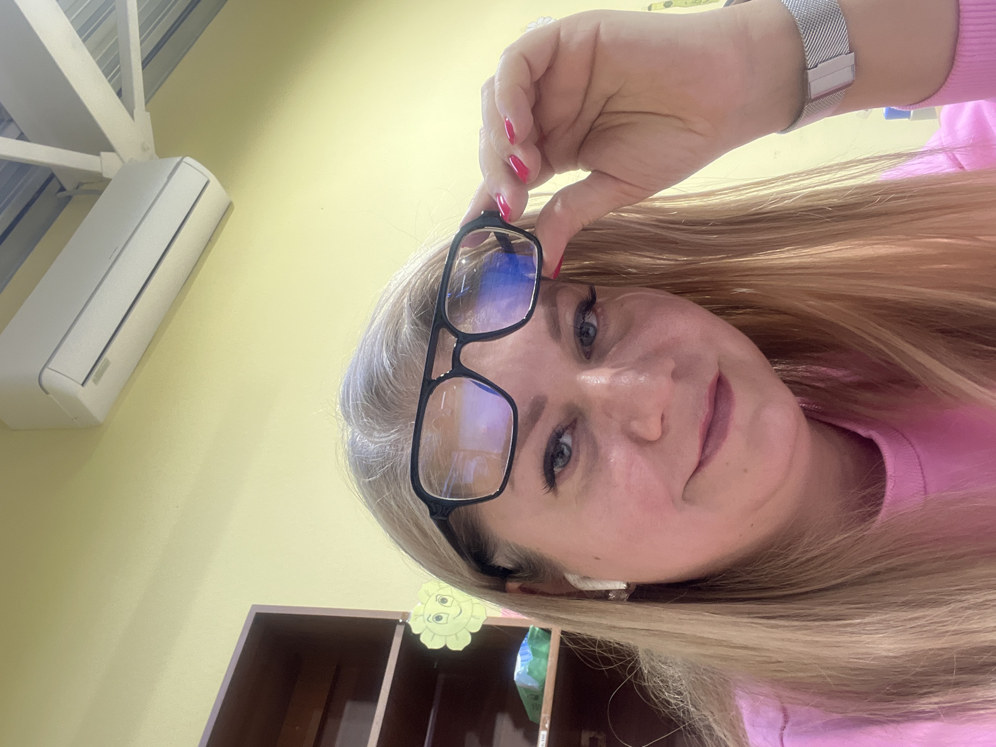 A fair-skinned woman with blonde hair holds black-framed glasses up to her face. She is wearing a pink sweater and a silver wristwatch. In the background is a light yellow wall with a white air conditioner mounted on it. A wooden cabinet with a yellow flower sticker is partially visible in the lower left corner. There is no information to suggest the location is in Izhevsk, Russia.