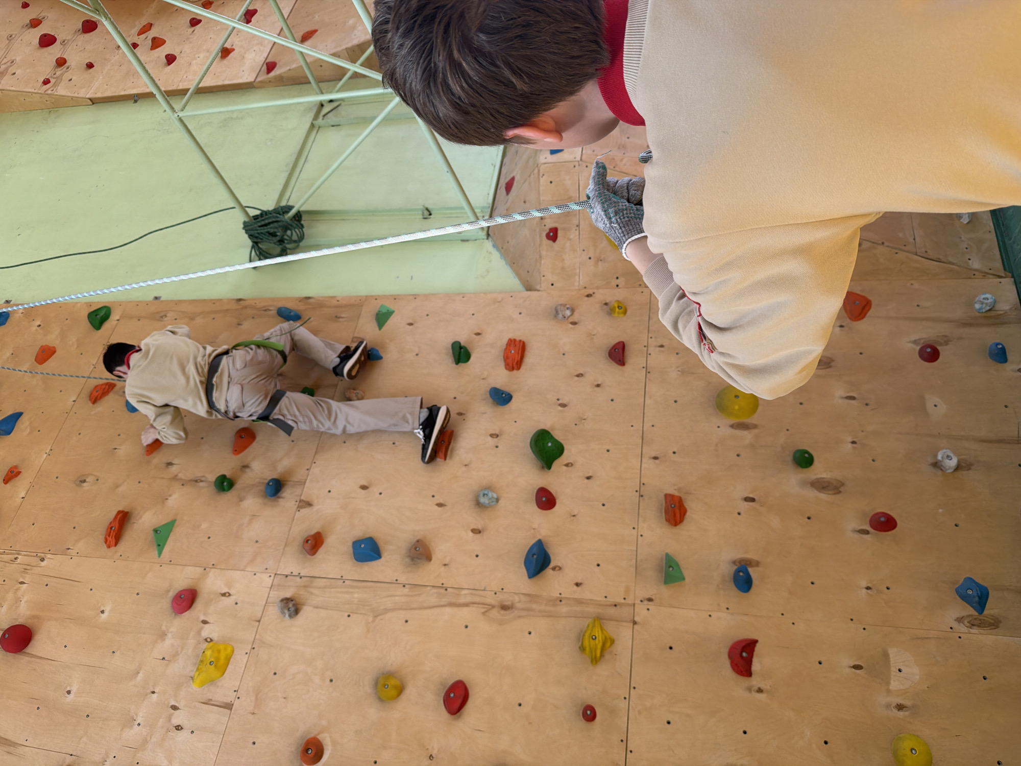A climbing wall is shown in the city of Izhevsk, Russia. A person in light colored clothing is climbing the wall, and another person is standing on the right, holding a rope. The climbing wall is made of plywood and has various colored holds attached to it. A green metal structure is visible on the ceiling with a rope looped around it.