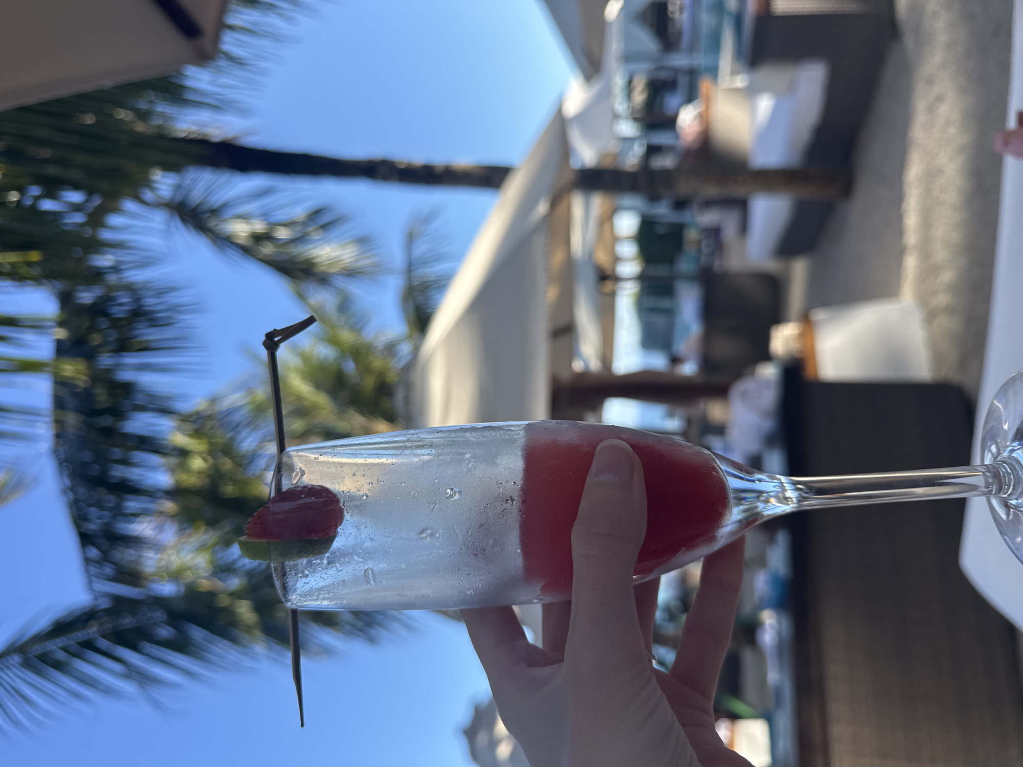 A hand holds a champagne flute filled with a red beverage and garnished with a strawberry slice and a lime wedge. The flute is positioned in the foreground against a backdrop of palm trees, clear blue sky, and resort lounge chairs with umbrellas. The scene appears to be outdoors, possibly at a tropical resort.