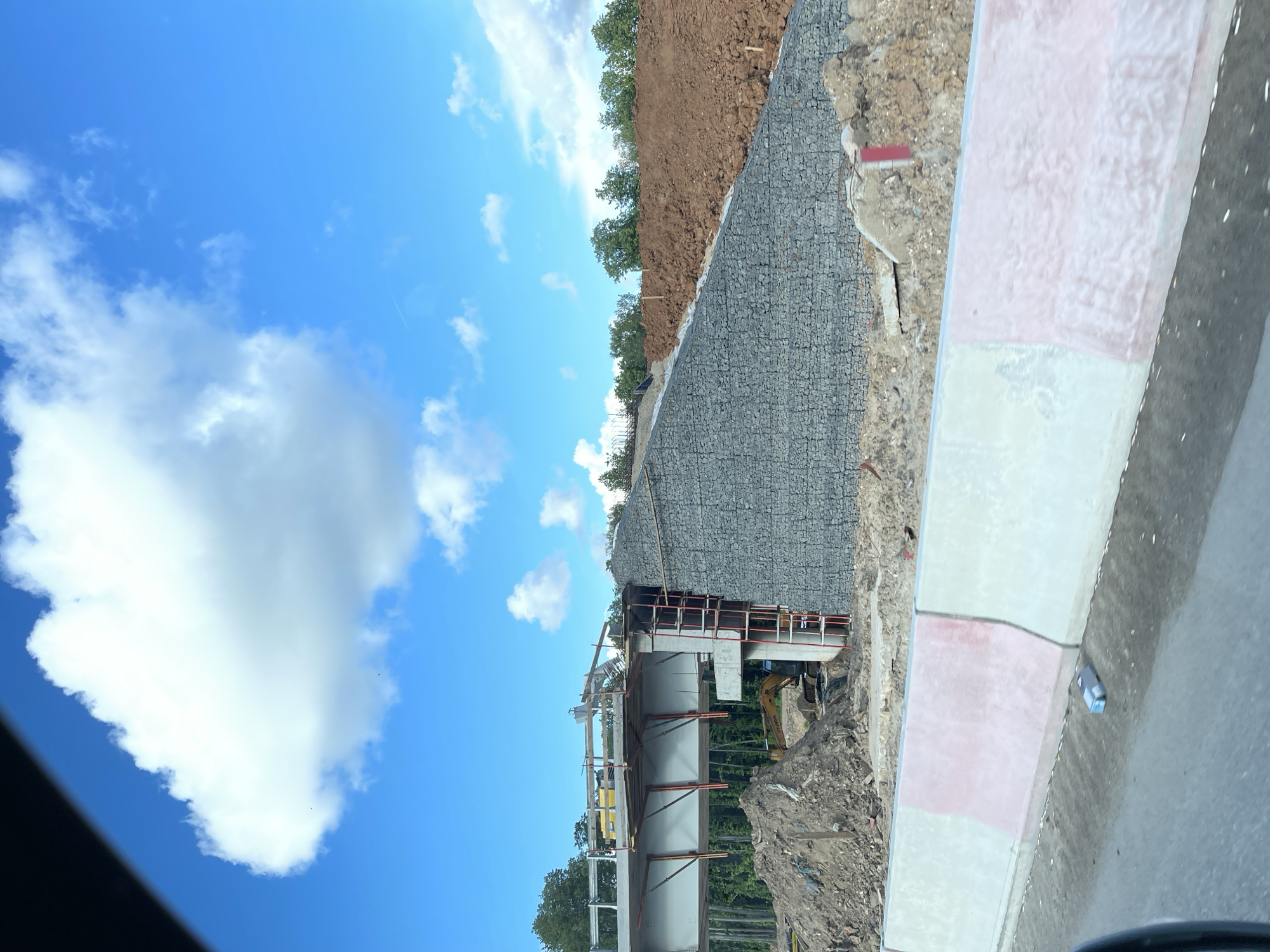 The image depicts a construction site in Novodrozhzhino, Russia. A newly constructed bridge is visible, with supporting steel and concrete structures. An excavator is parked nearby, surrounded by dirt and construction debris. Adjacent to the construction site is a retaining wall made of gabions, preventing soil erosion. The sky is blue with scattered clouds. A segment of a roadside curb with painted white and pink sections is present in the foreground.