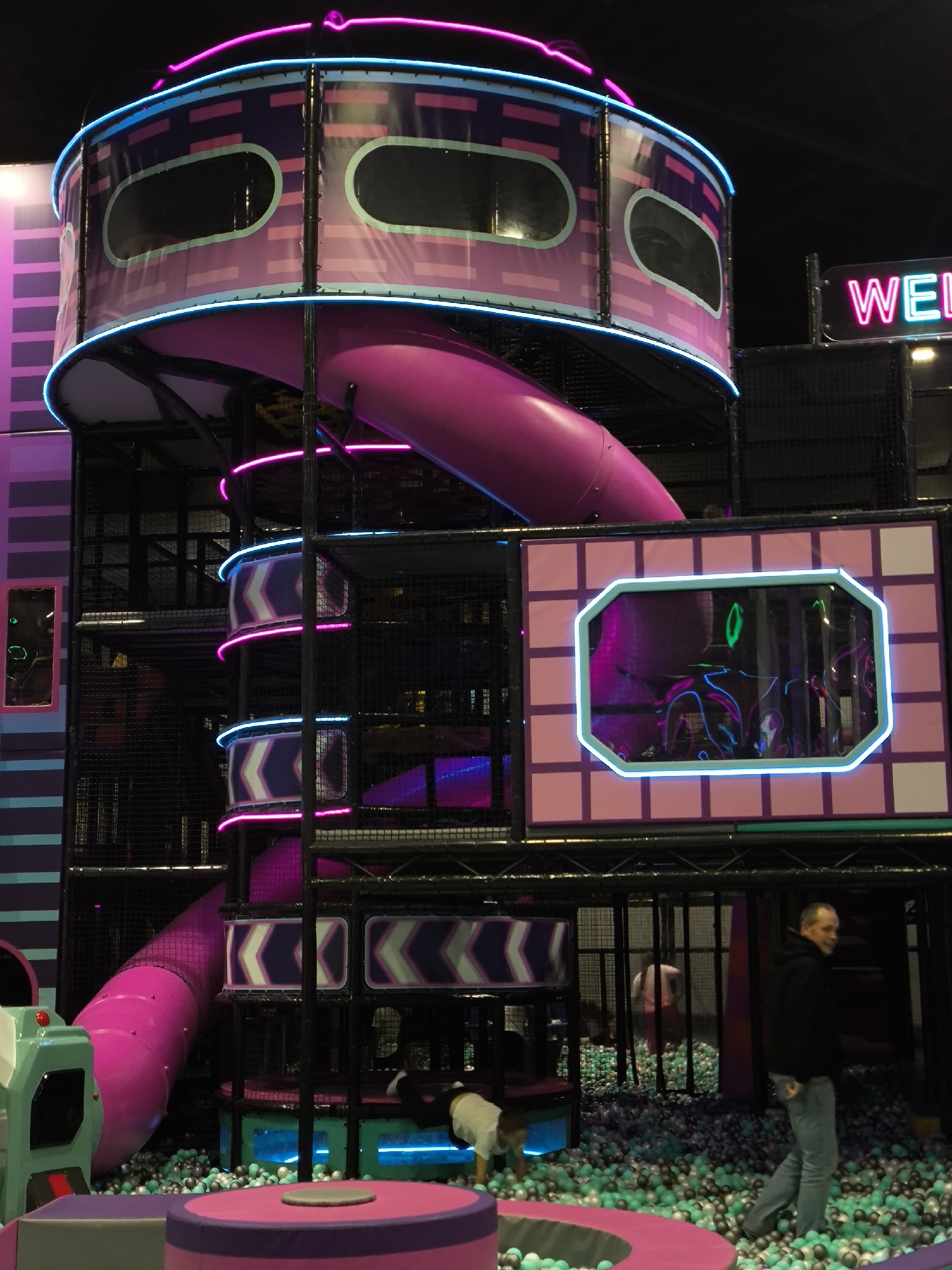 The image shows a brightly lit indoor children's play area, likely an amusement center. Dominating the scene is a multi-level play structure with a large, curved pink slide. The structure is adorned with neon pink and blue lights and features purple and pink geometric designs. There are also several windows or observation areas on the structure. In the background, a "WELCOME" sign with neon letters is visible. The floor of the play area is covered in a ball pit filled with green and grey balls. A man in a black hoodie and jeans is walking on the right side of the image, and a child in a white shirt is bent over in the ball pit on the left. Another person, possibly a child, is partially visible in the background through a caged section. The overall environment suggests a playful and energetic atmosphere. There is no information in the image that indicates the location is in Орловка, Russia.
